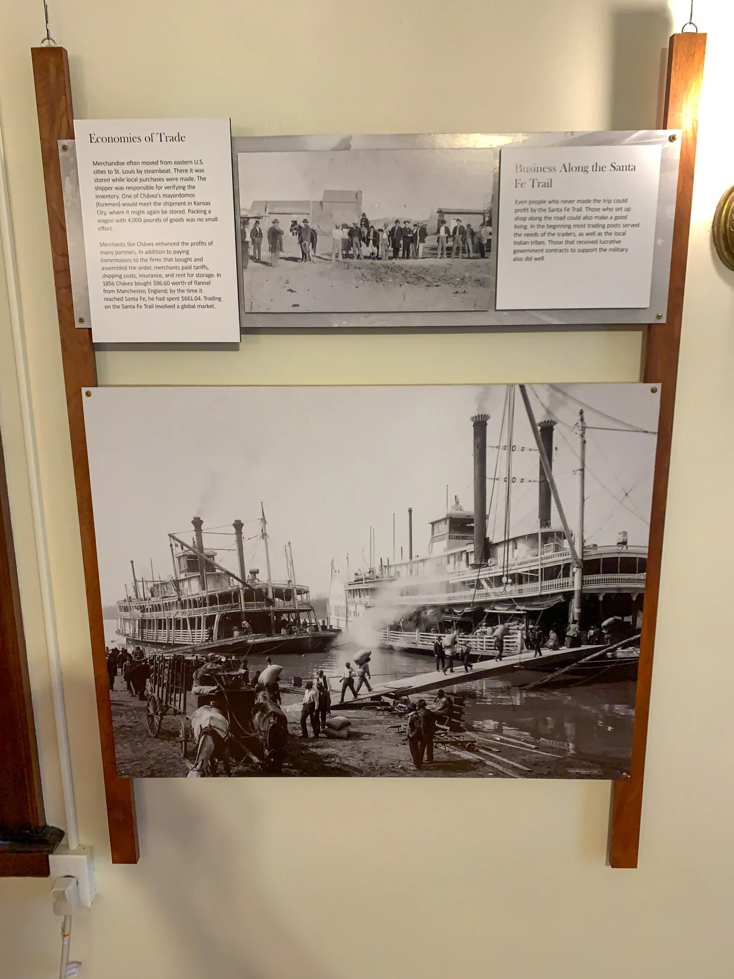 Museum display featuring historical photographs. The top section describes "Economies of Trade" and "Business Along the Santa Fe Trail," with a small image. Below is a large black-and-white photo of people and steamboats at a busy river dock, depicting 19th-century commerce.