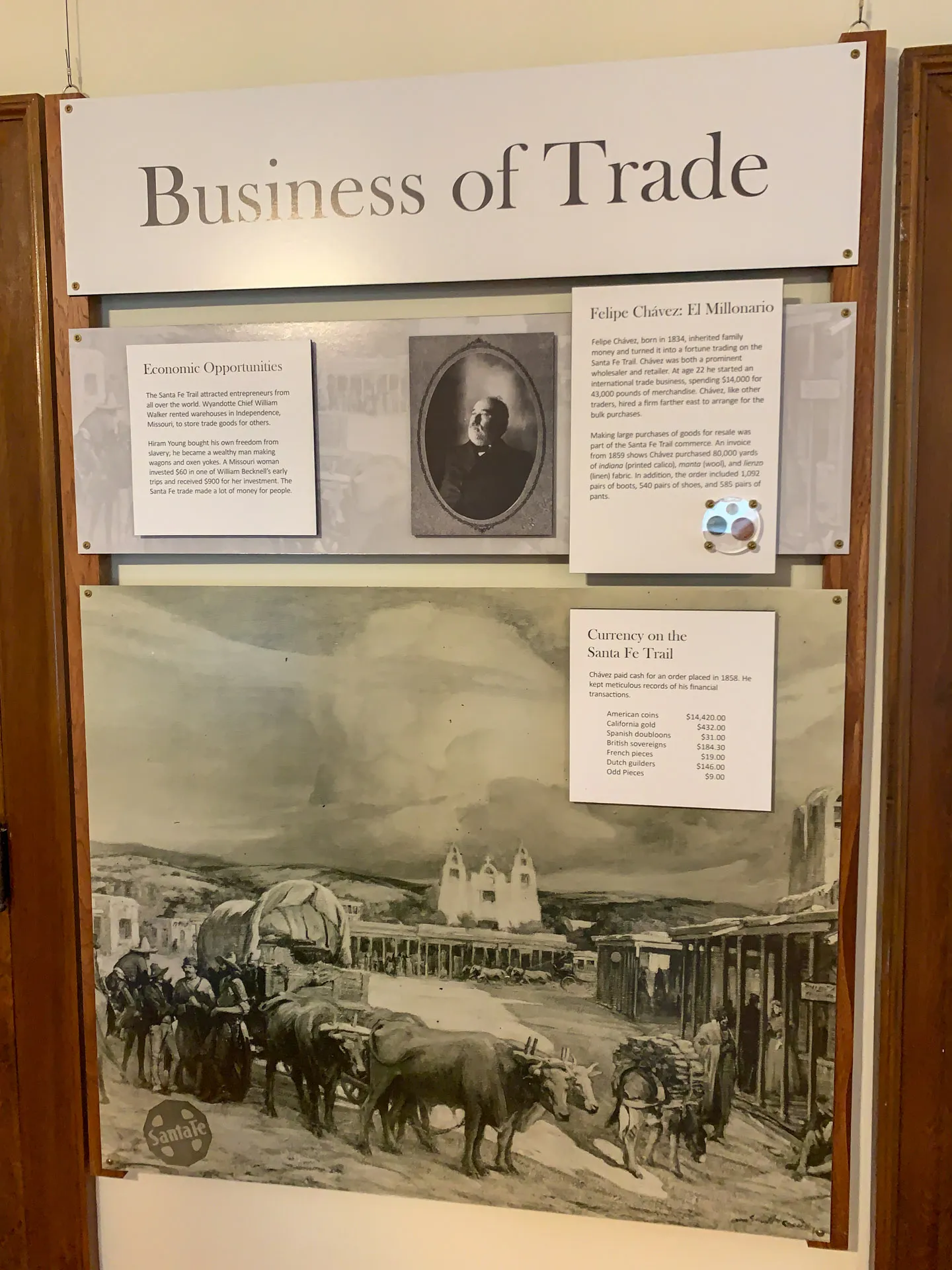 A historical display titled "Business of Trade" features three panels. The top panel discusses economic opportunities, the middle shows a portrait of Felipe Chávez with a biography, and the bottom displays a historic trade scene with oxen and carts near buildings labeled "Santa Fe.