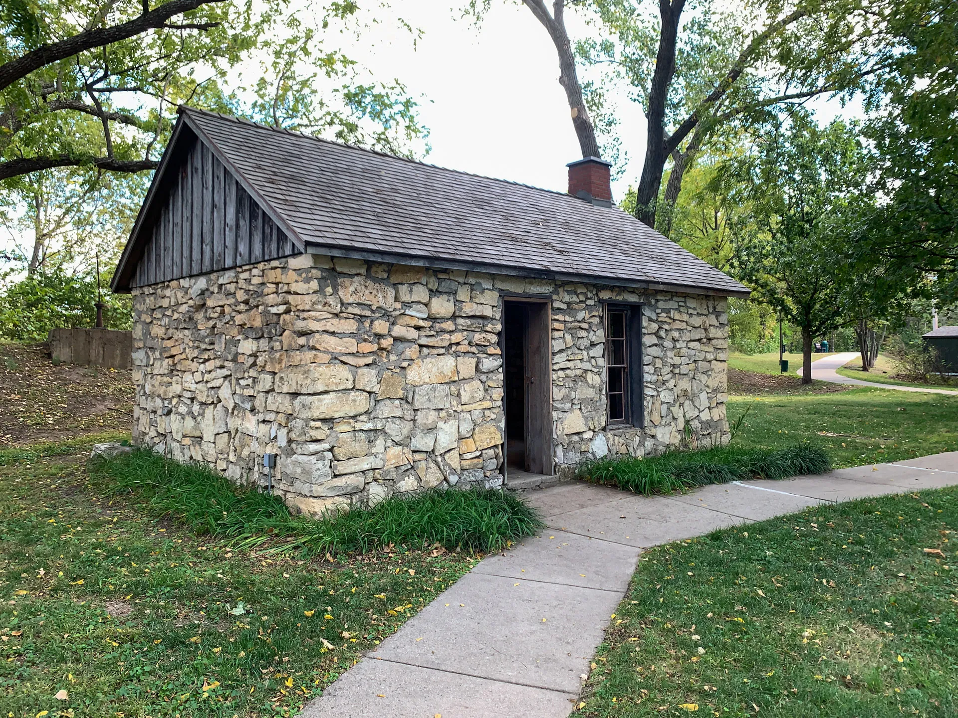 A small stone cabin with a wooden gabled roof stands in a grassy area. The cabin has a chimney, two windows with dark frames, and a wooden door. A concrete pathway leads to the entrance. Tall trees surround the building, with some leaves scattered on the grass.