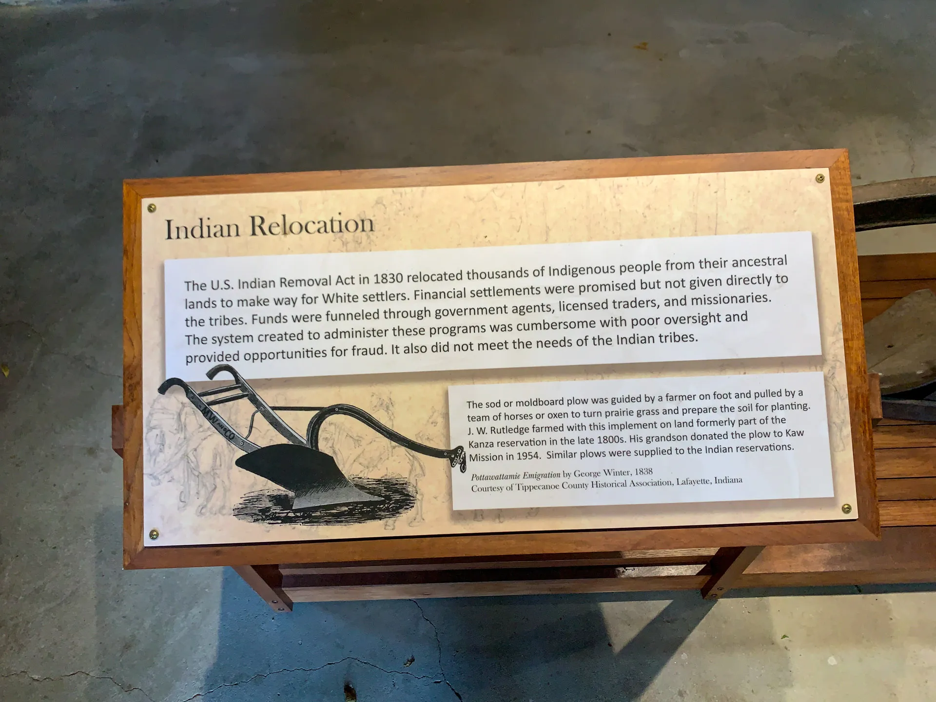 A wooden display featuring an informational plaque titled "Indian Relocation." The plaque describes the 1830 Indian Removal Act, noting relocation issues, financial settlements, and governmental mismanagement. Below is an illustration of a traditional plow. A bench is partly visible.