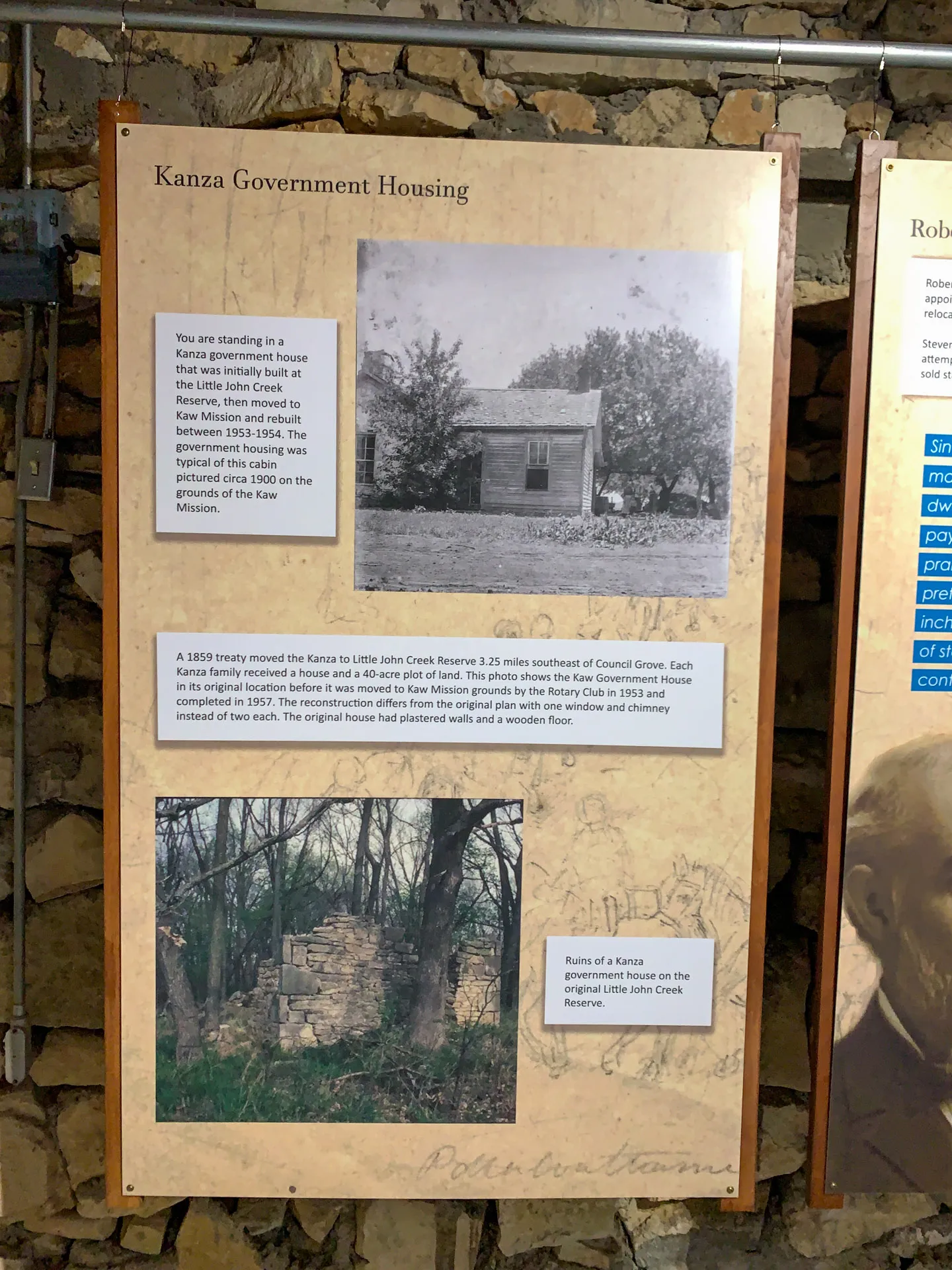 A museum display board titled "Kanza Government Housing" features two black and white photos. The top image shows a 19th-century house with a wooden fence. The bottom image depicts ruins of a stone structure. Text describes the history of Kanza housing on the Little John Creek Reserve.
