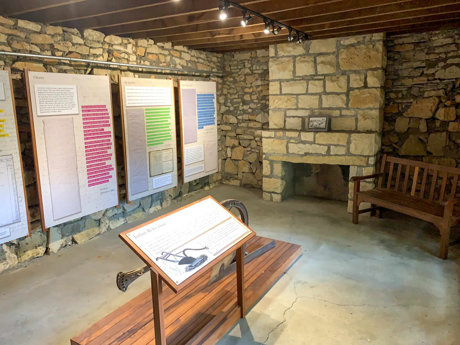 A museum exhibit features several large text panels with historical information displayed on stone walls. A wooden bench sits on the right, and a central stand holds an artifact with descriptive text. The room has a rustic stone aesthetic with overhead lighting.