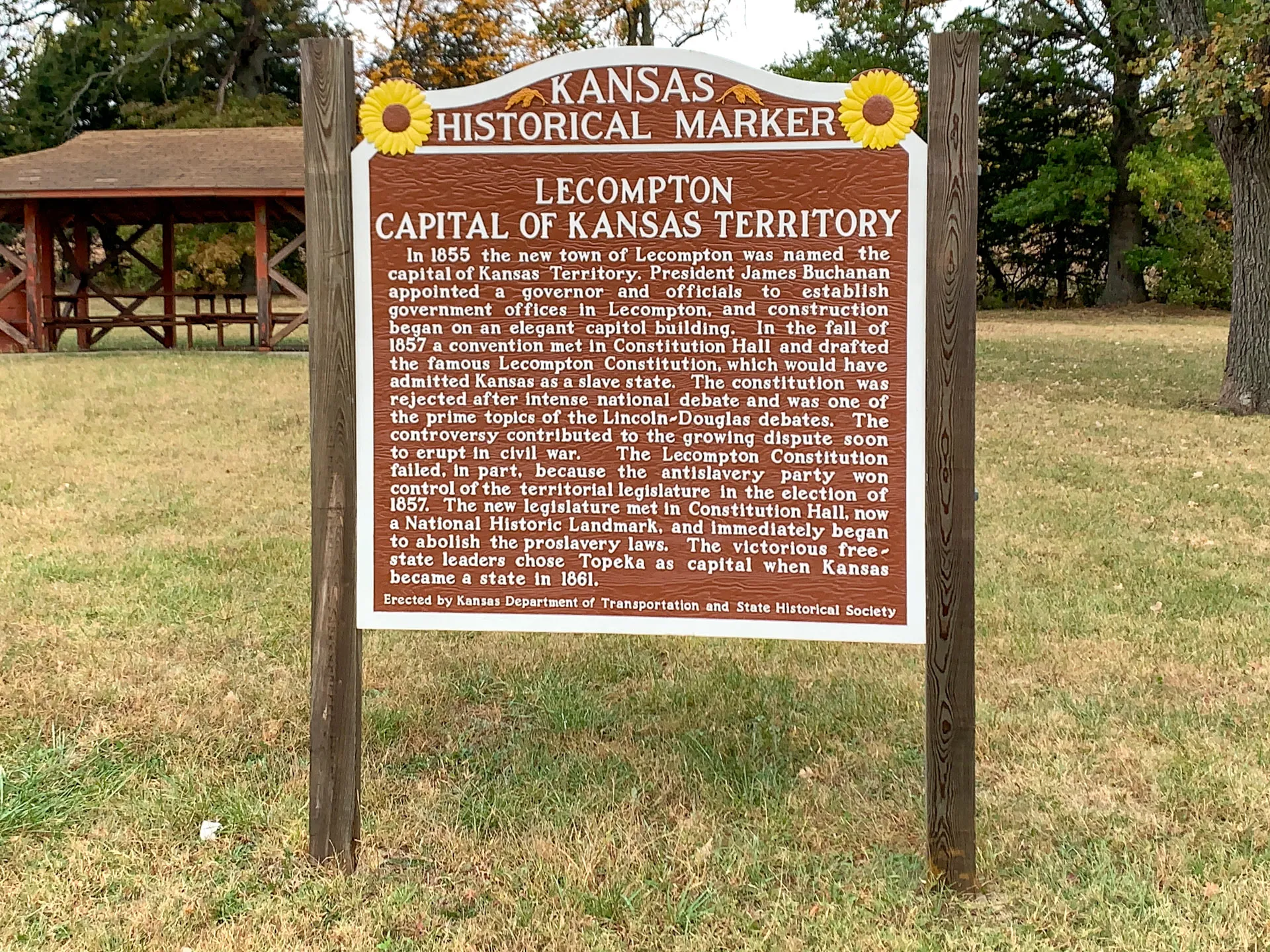 Kansas historical marker titled "Lecompton Capital of Kansas Territory" with text about its founding, constitutional convention, and legislative history. A pavilion and trees are in the background.