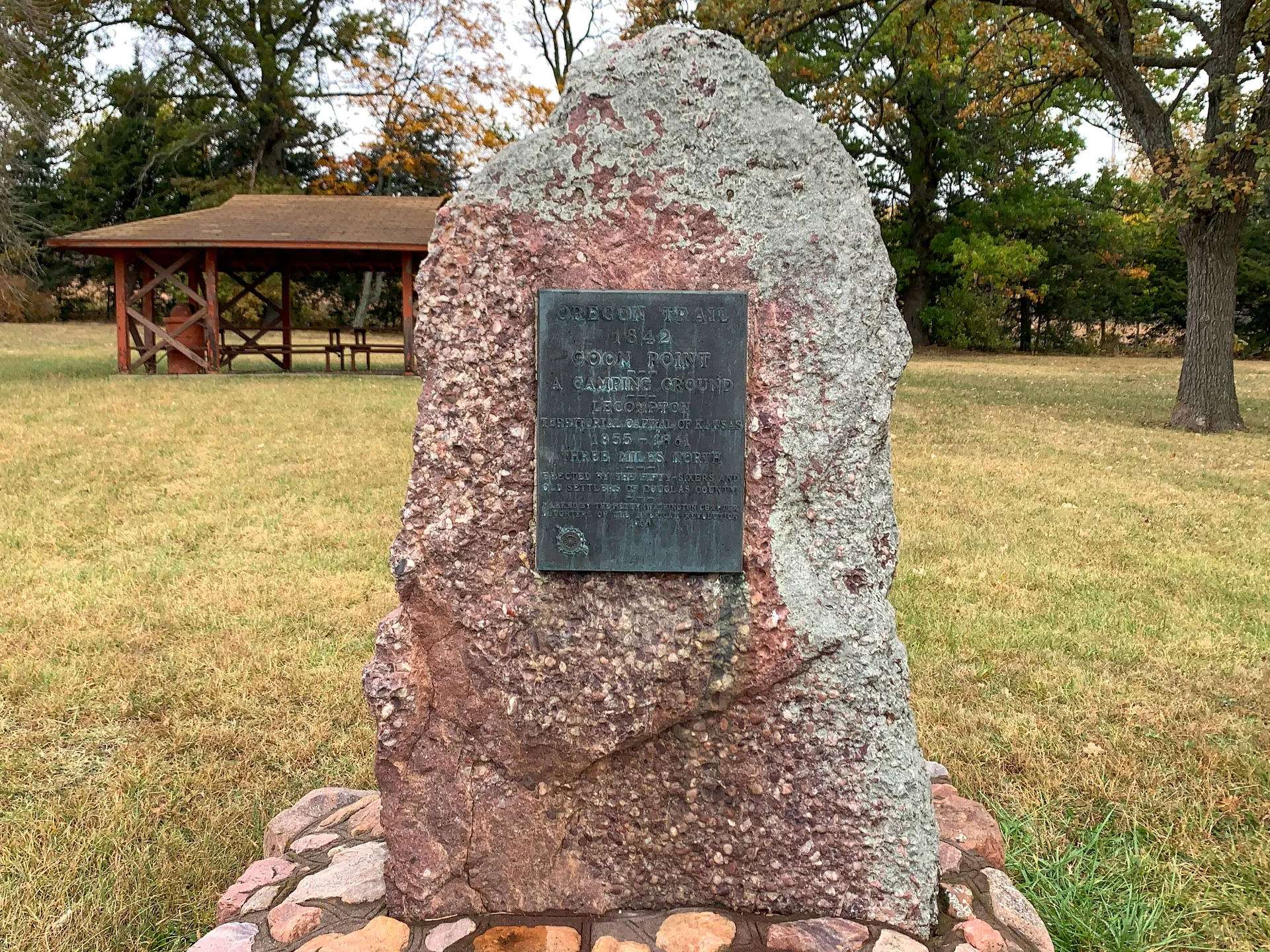 Stone monument with a plaque stands on grassy ground, surrounded by trees and near a wooden pavilion. Plaque text is not fully visible.