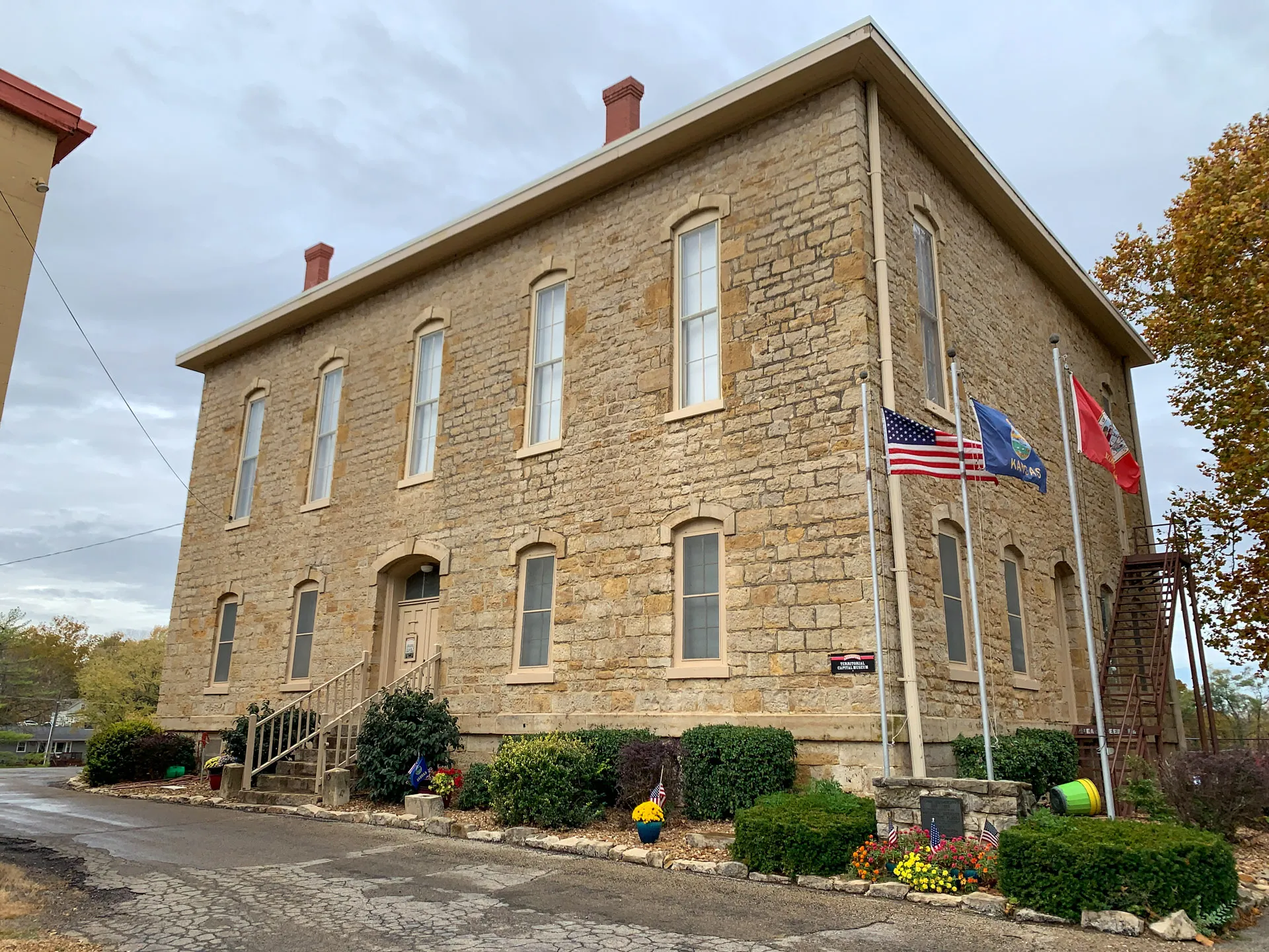 A two-story stone building with tall windows, three flags in the front, and a small garden. Overcast sky in the background.