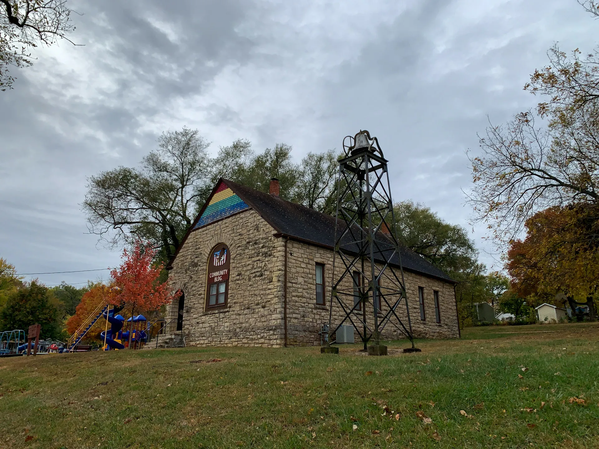 A small stone building with a gable roof stands near a metal tower. The building and nearby trees are surrounded by a grassy field. A playground is visible to the left. Cloudy sky overhead.