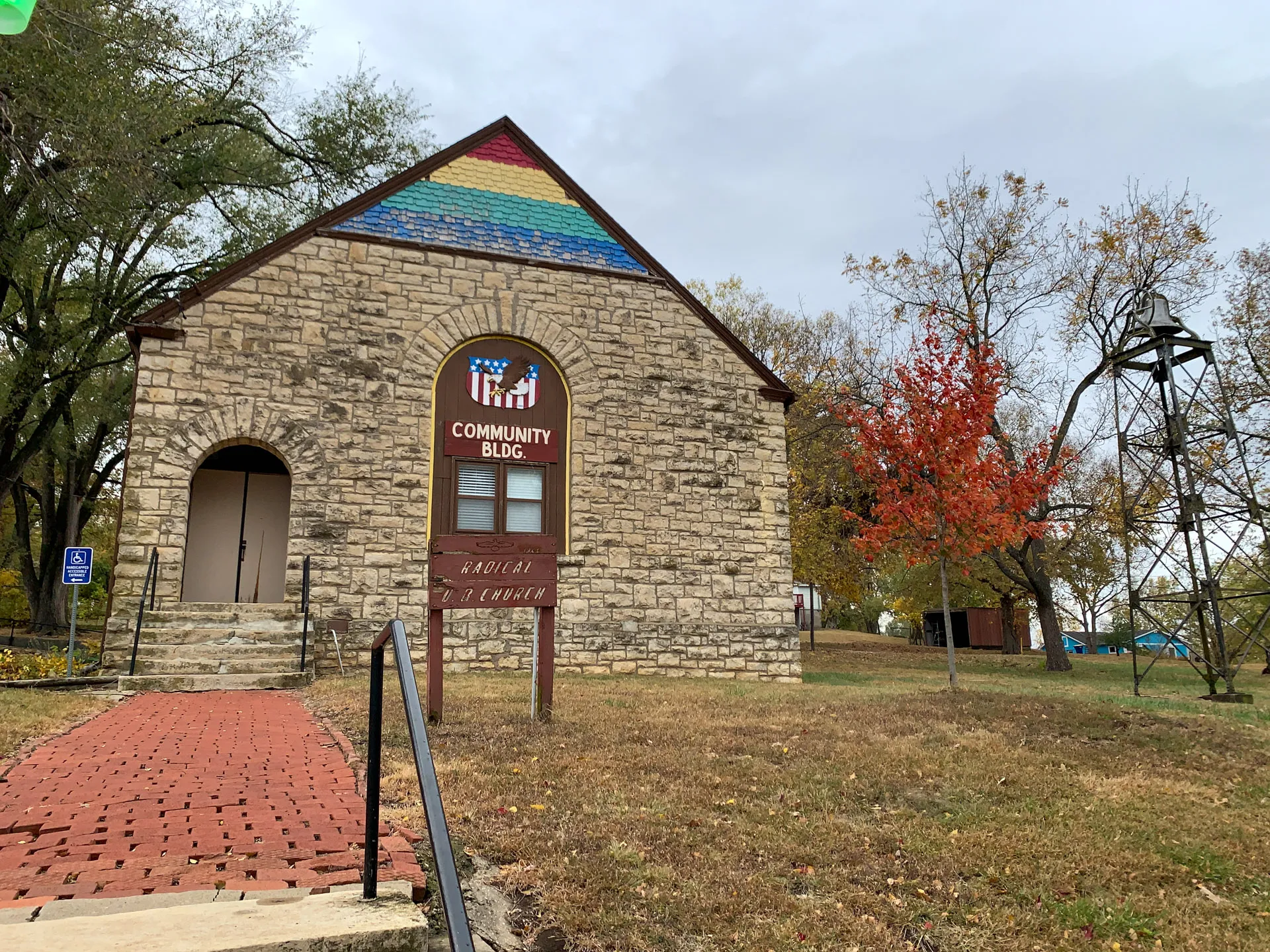 Stone building labeled "Community Bldg" with a rainbow-painted roof section, surrounded by trees and a brick pathway.