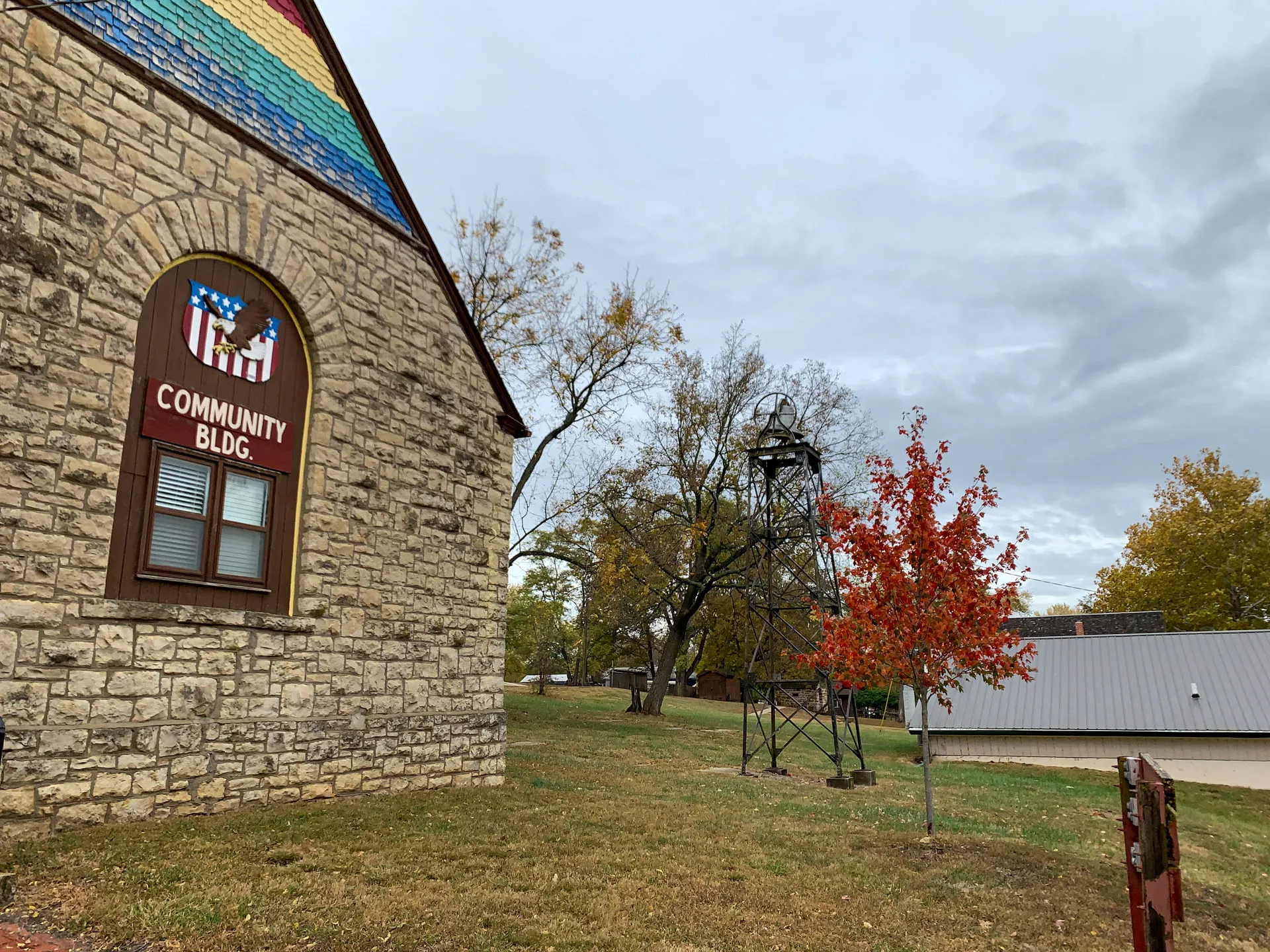 Stone building with "Community Bldg" sign and rainbow roof section; nearby, a small metal tower stands beside a red-leaved tree.