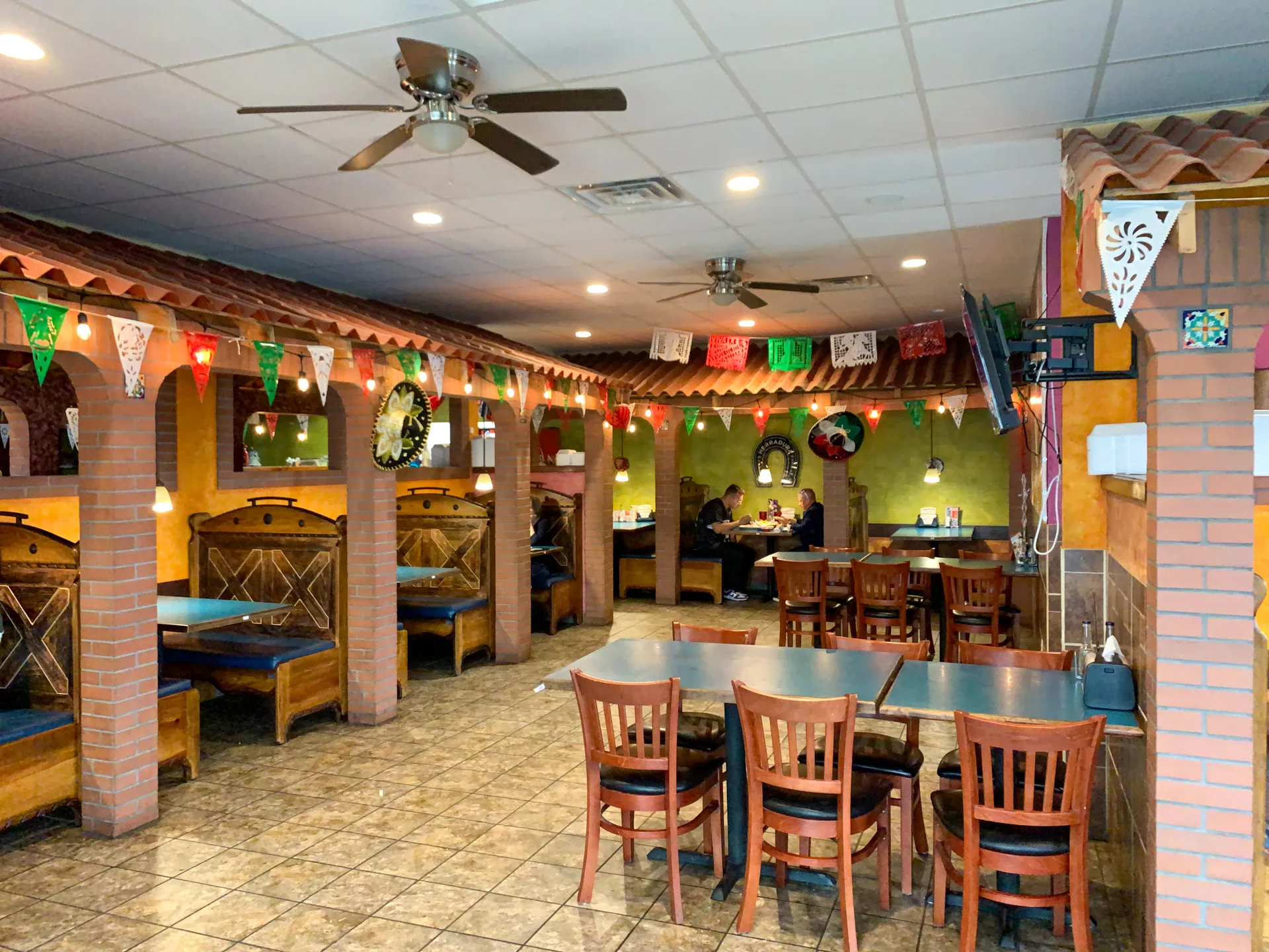 A cozy restaurant interior with brick arches and warm lighting. Wooden booths line the left side, with empty tables and chairs in the center. Colorful string flags hang from the ceiling. The walls are adorned with festive decorations, and ceiling fans are visible above.