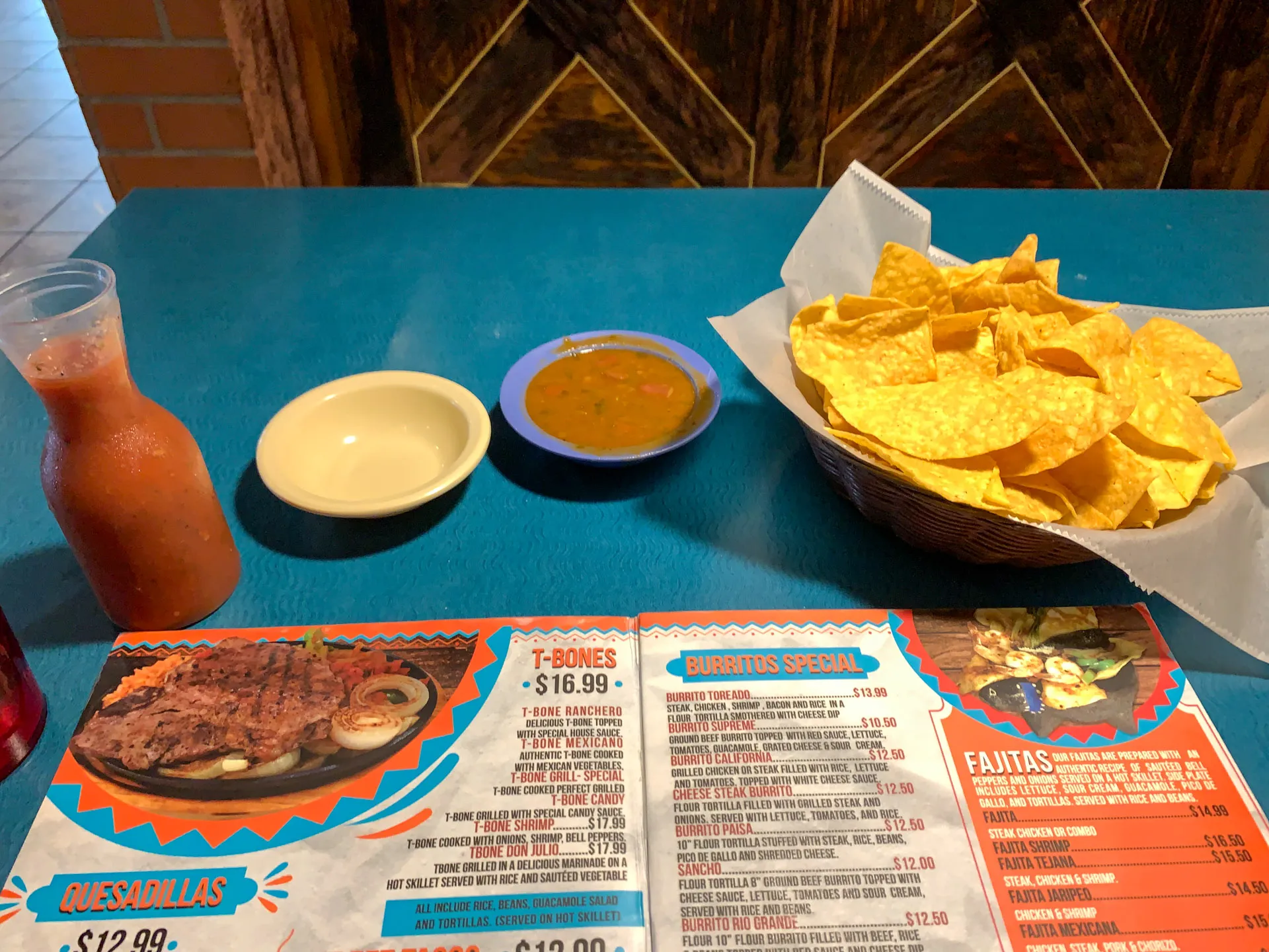 A table at a Mexican restaurant features a bright menu open to pages showing various dishes like quesadillas and burritos. A basket of tortilla chips, a bowl of salsa, and a small bottle of hot sauce sit on the blue tabletop. The background includes a wooden partition.