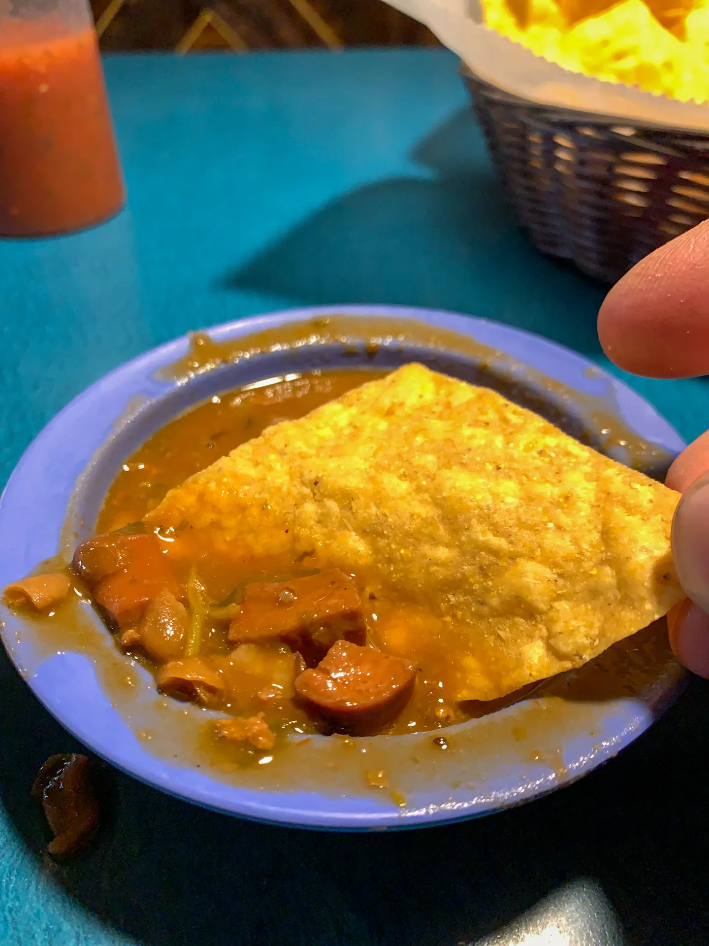 A hand dipping a tortilla chip into a bowl of chili con carne on a blue plate. The chili contains beans and chunks of meat in a rich sauce. In the background, there is a basket of tortilla chips and a bottle of red sauce on a teal table.