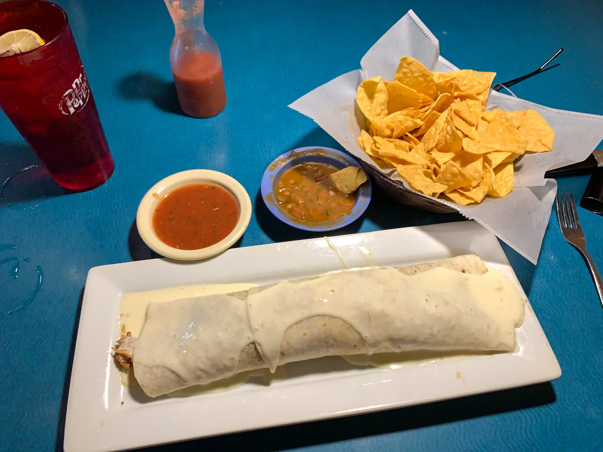 A burrito covered in white cheese sauce sits on a rectangular plate. Nearby are bowls of red and green salsa and a basket of tortilla chips on a blue table. A bottle of hot sauce and a tall red glass filled with a drink and a lemon slice are in the background, next to a fork.