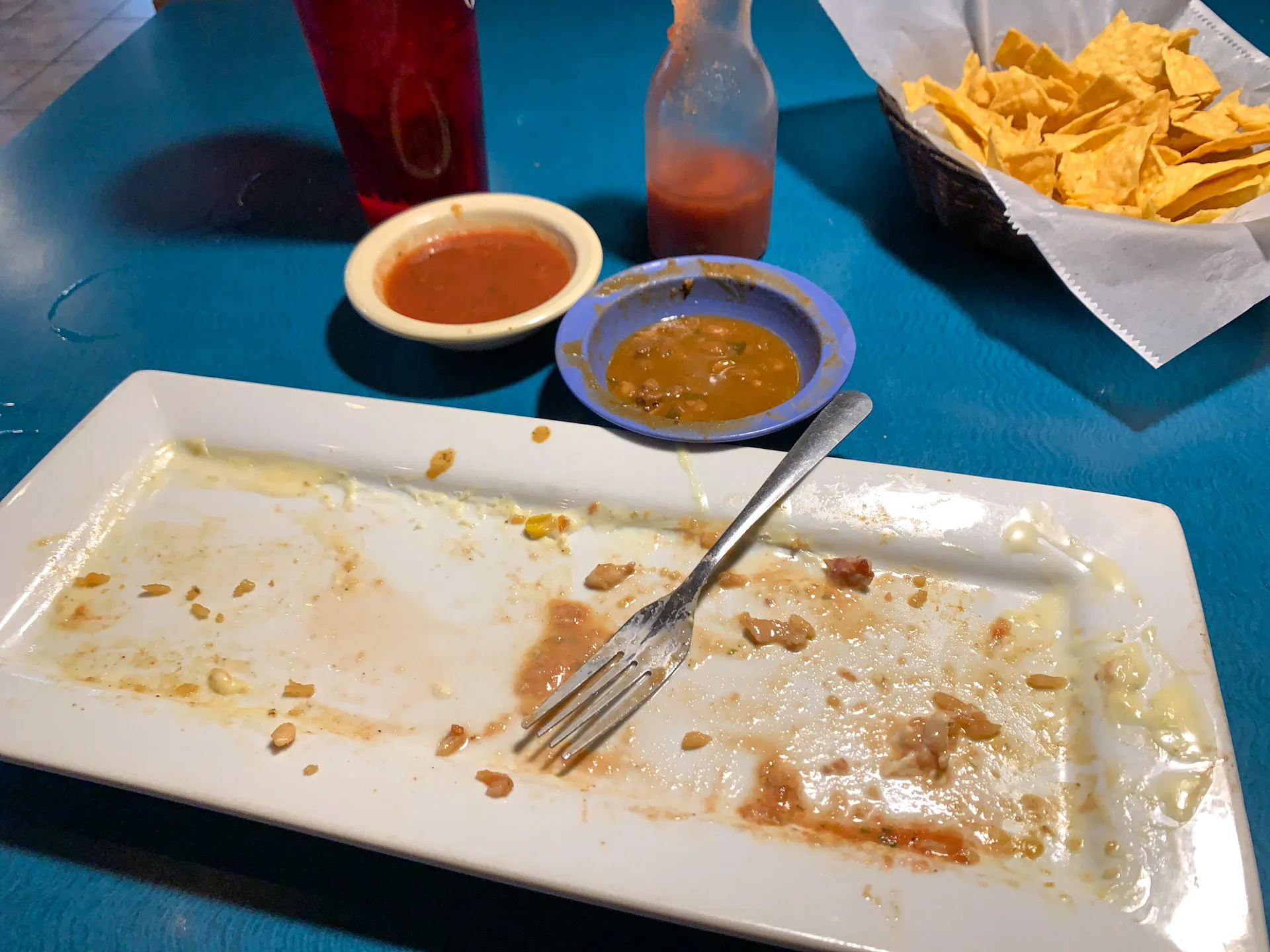 An empty rectangular plate with food remnants and a fork sits on a blue table. Nearby are two small bowls: one with red sauce, the other with green. A bottle of hot sauce and a basket of tortilla chips are beside a red plastic cup.