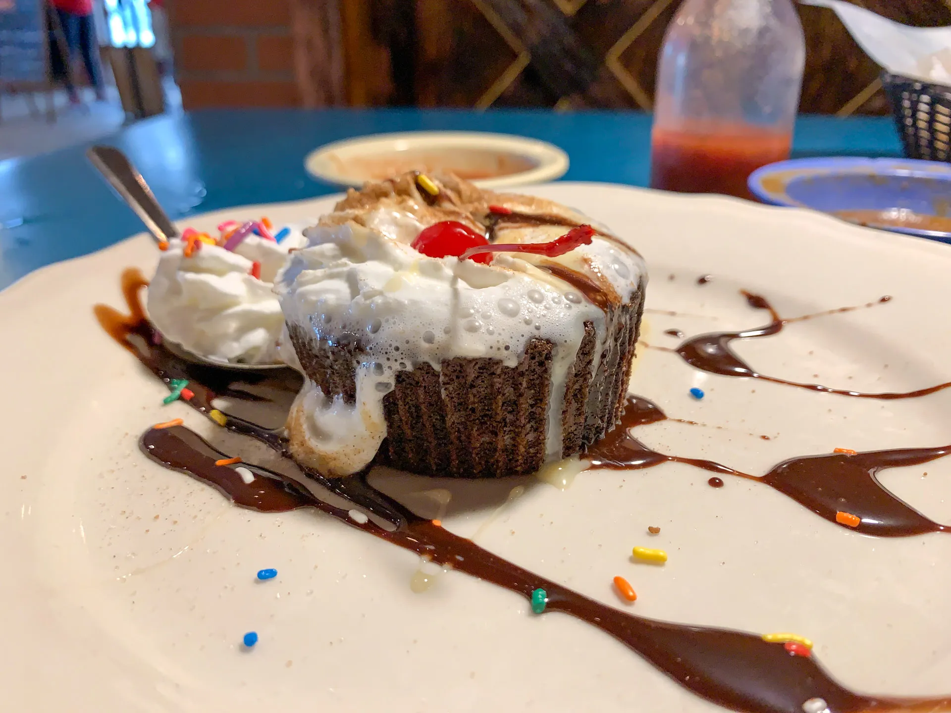A chocolate dessert with whipped cream, a cherry on top, and multicolored sprinkles is on a white plate. Chocolate sauce is drizzled on the plate. A spoon is on the dessert. In the background, a bottle of red sauce and a blue table are visible.