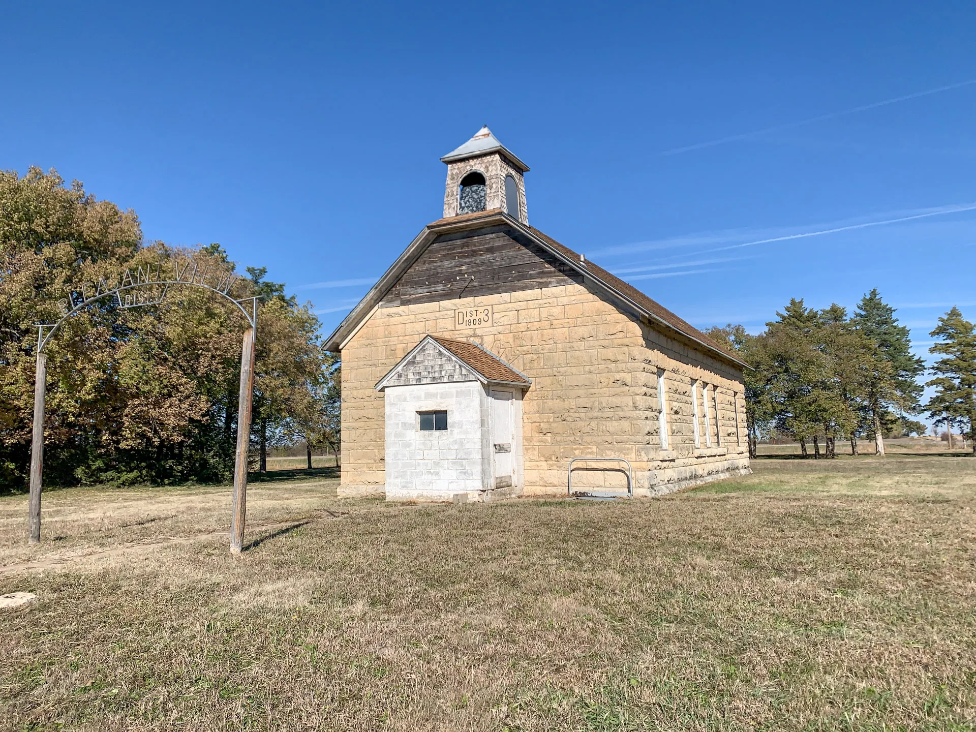 A historic, small stone schoolhouse with a gray roof and a simple bell tower stands in an open grassy field under a clear blue sky. An arched metal sign on the left reads "BEN HALL." Surrounding trees add to the serene rural setting.