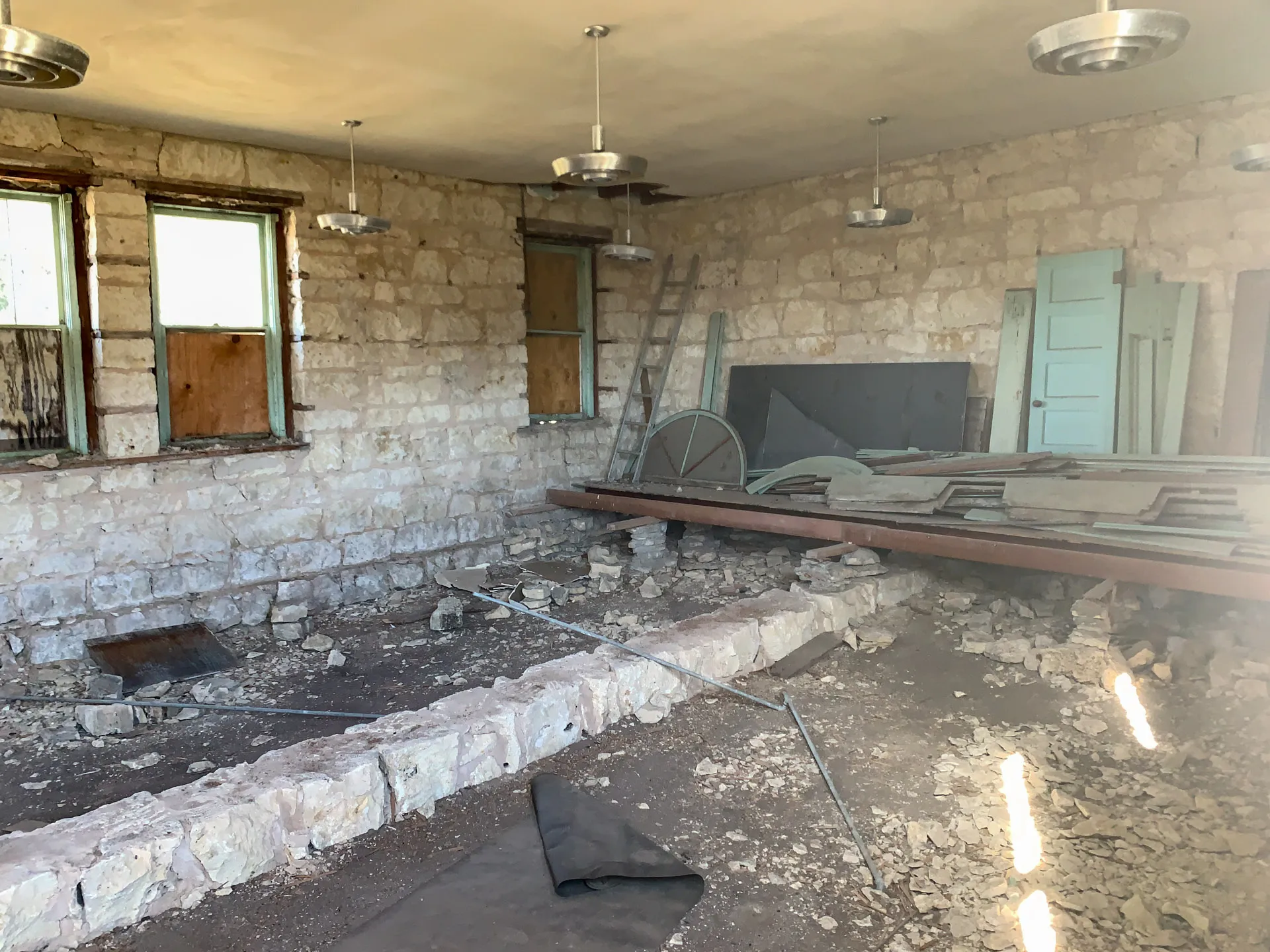 An abandoned stone room with exposed beam ceiling, featuring old-fashioned circular ceiling lights. The floor is partially collapsed, exposing rubble and debris. Leaning against the back wall are old doors, wooden planks, and a metal ladder. Two boarded windows are on the left wall.