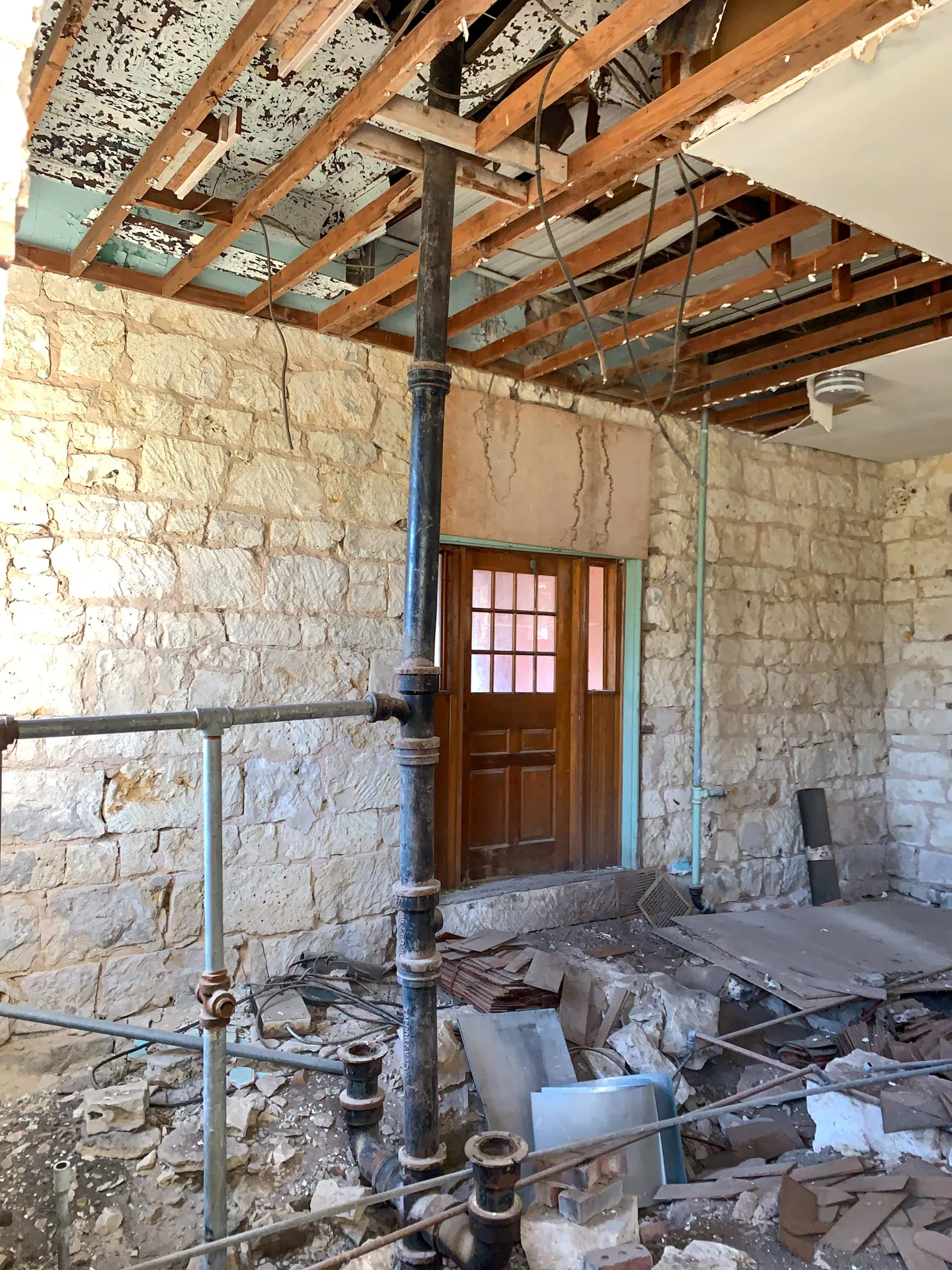 An old building interior with stone walls and exposed ceiling beams. A wooden door with a small glass window is centered. Metal pipes run vertically and horizontally. The floor is littered with debris, including pieces of drywall and wood. Wiring hangs from the ceiling.