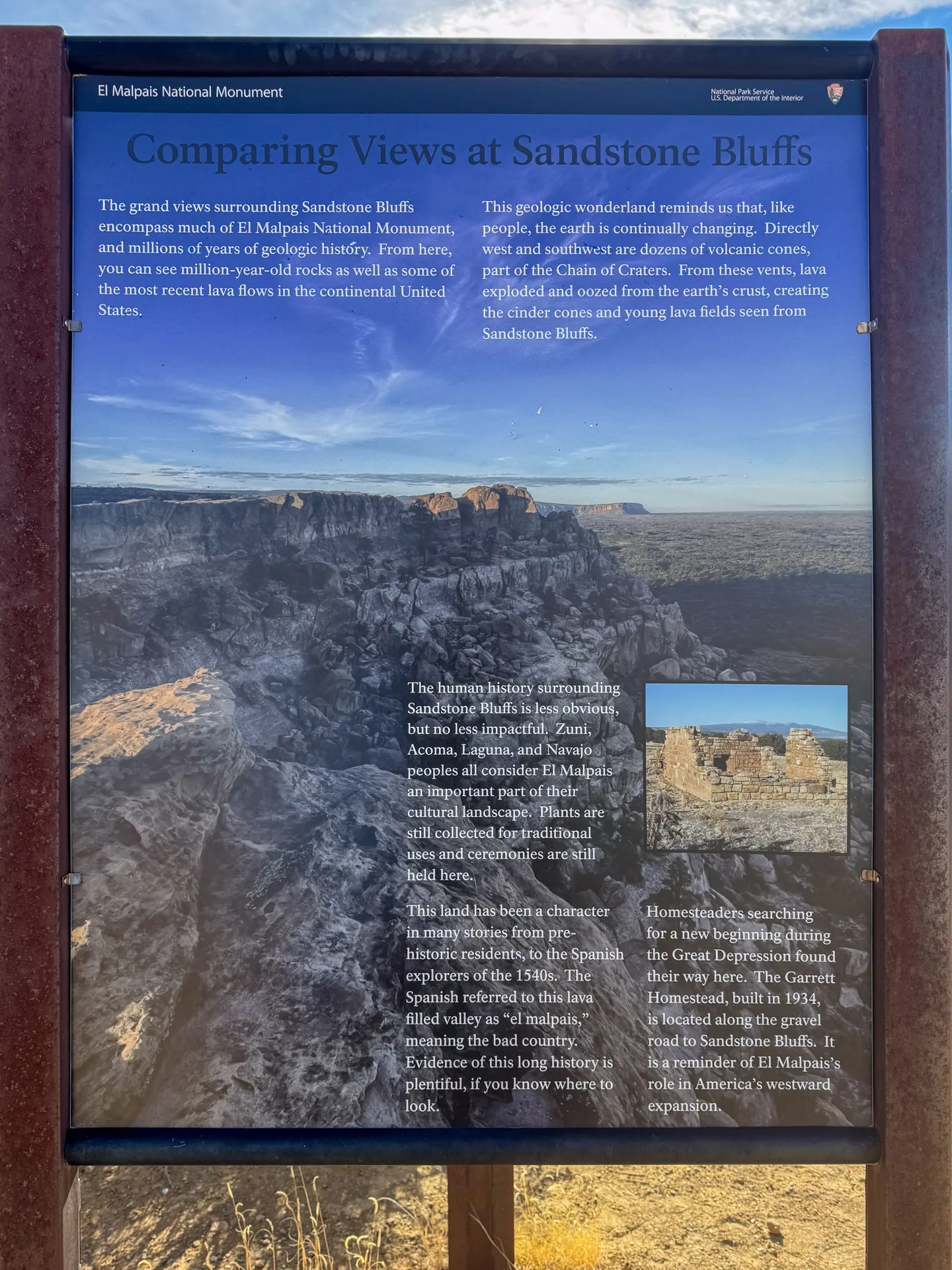 The image shows an informational sign at Sandstone Bluffs in El Malpais National Monument. It includes text on geological features and history, with a large photo of rugged bluffs and lava fields. A small inset photo shows eroded rock formations. The sky is clear and blue.