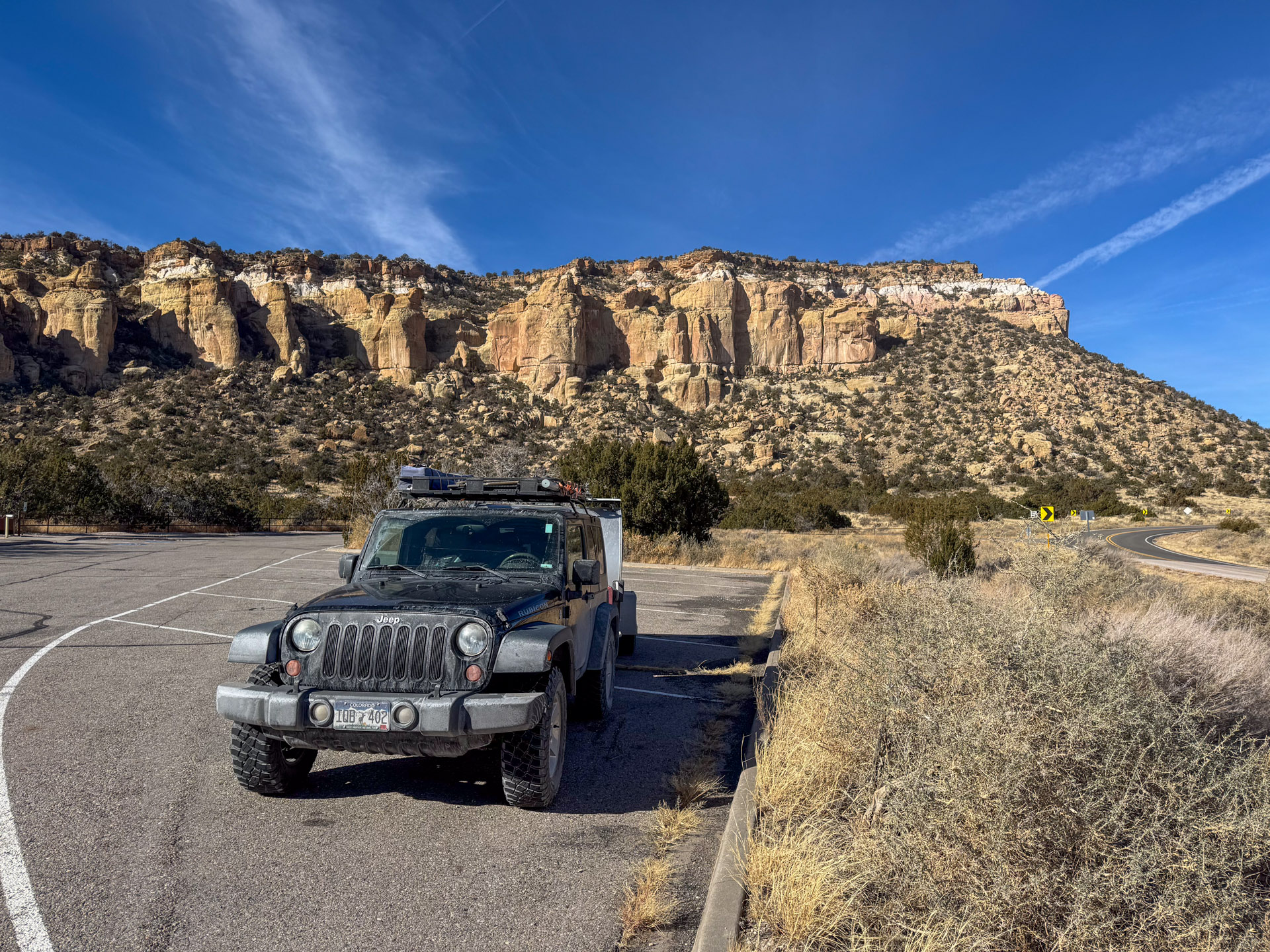 A rugged off-road vehicle is parked in an empty roadside area with desert shrubs. Behind, a towering rock formation under a clear blue sky creates a striking desert landscape. A winding road curves to the right, leading further into the desert environment.