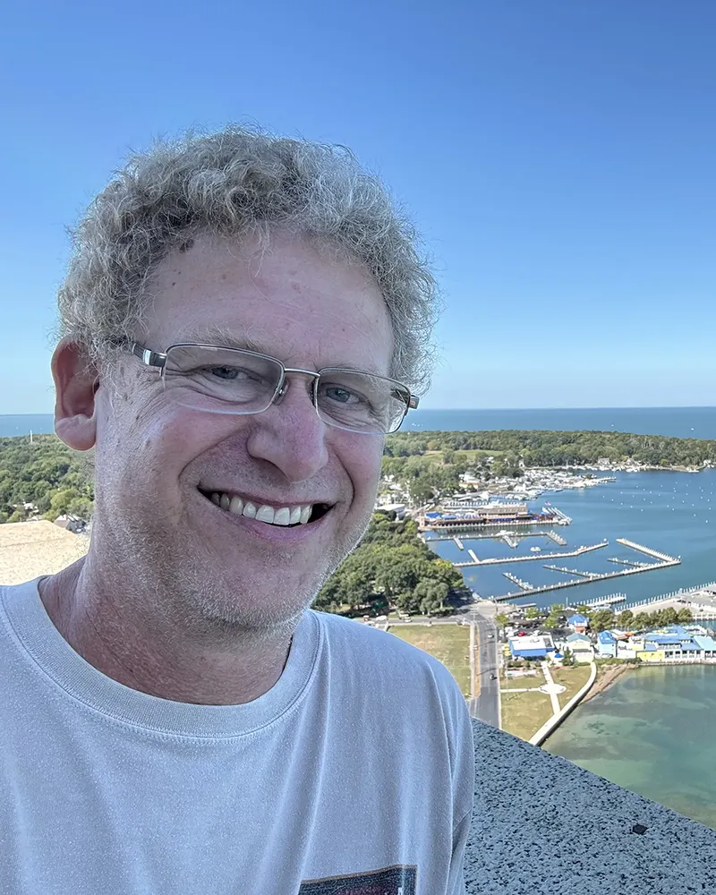 A man with curly hair and glasses smiles while taking a selfie. He is wearing a white t-shirt. Behind him, there is a panoramic view of a marina with numerous boats, surrounded by green trees. The sky is clear and blue.