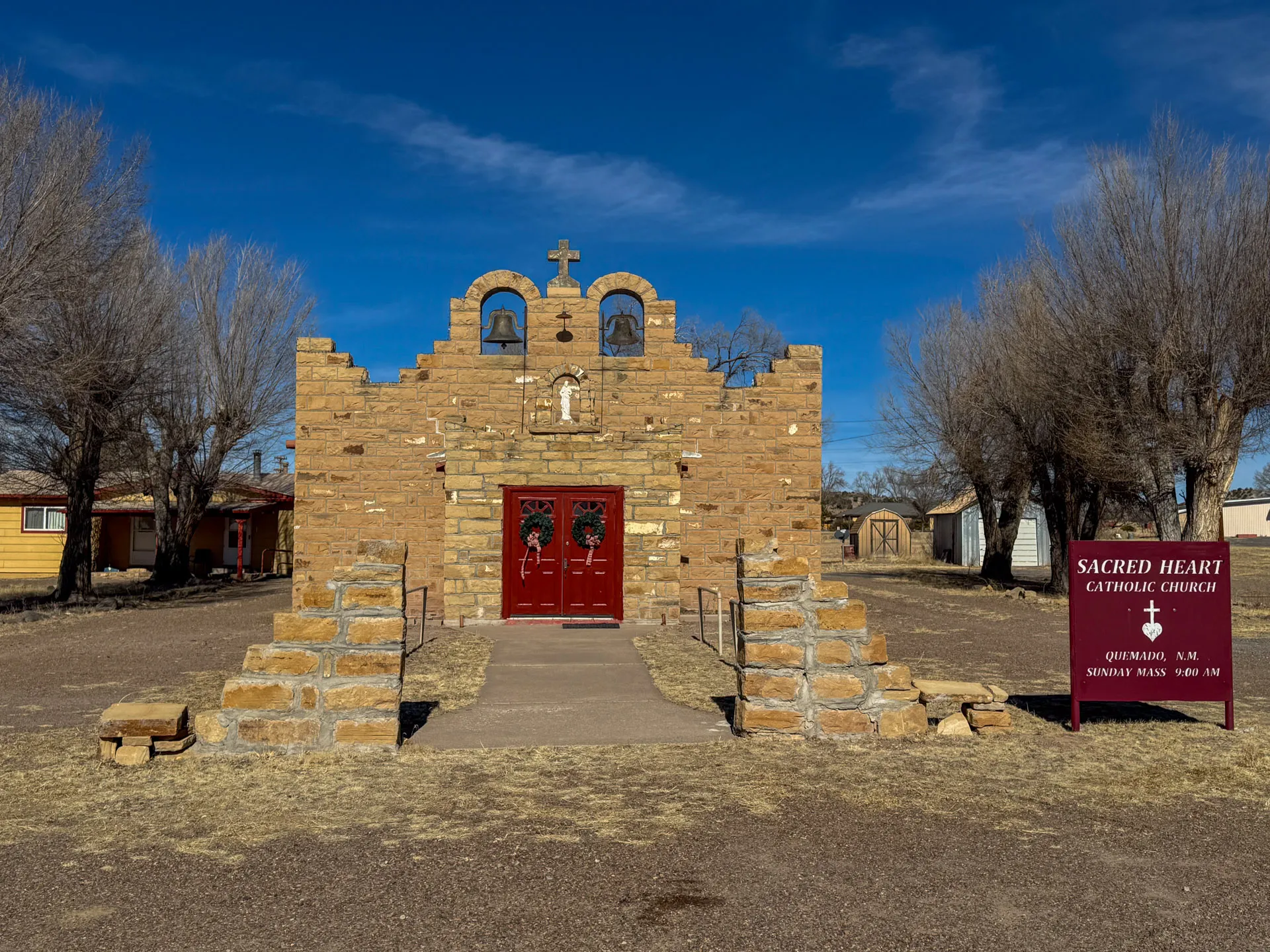 Historic stone church with uneven brick facade, red double doors, and twin bell arches. A cross adorns the top. Leafless trees flank the structure. A sign reads: "Sacred Heart Catholic Church, Quemado, N.M., Sunday Mass 8:00 AM." Clear blue sky overhead.
