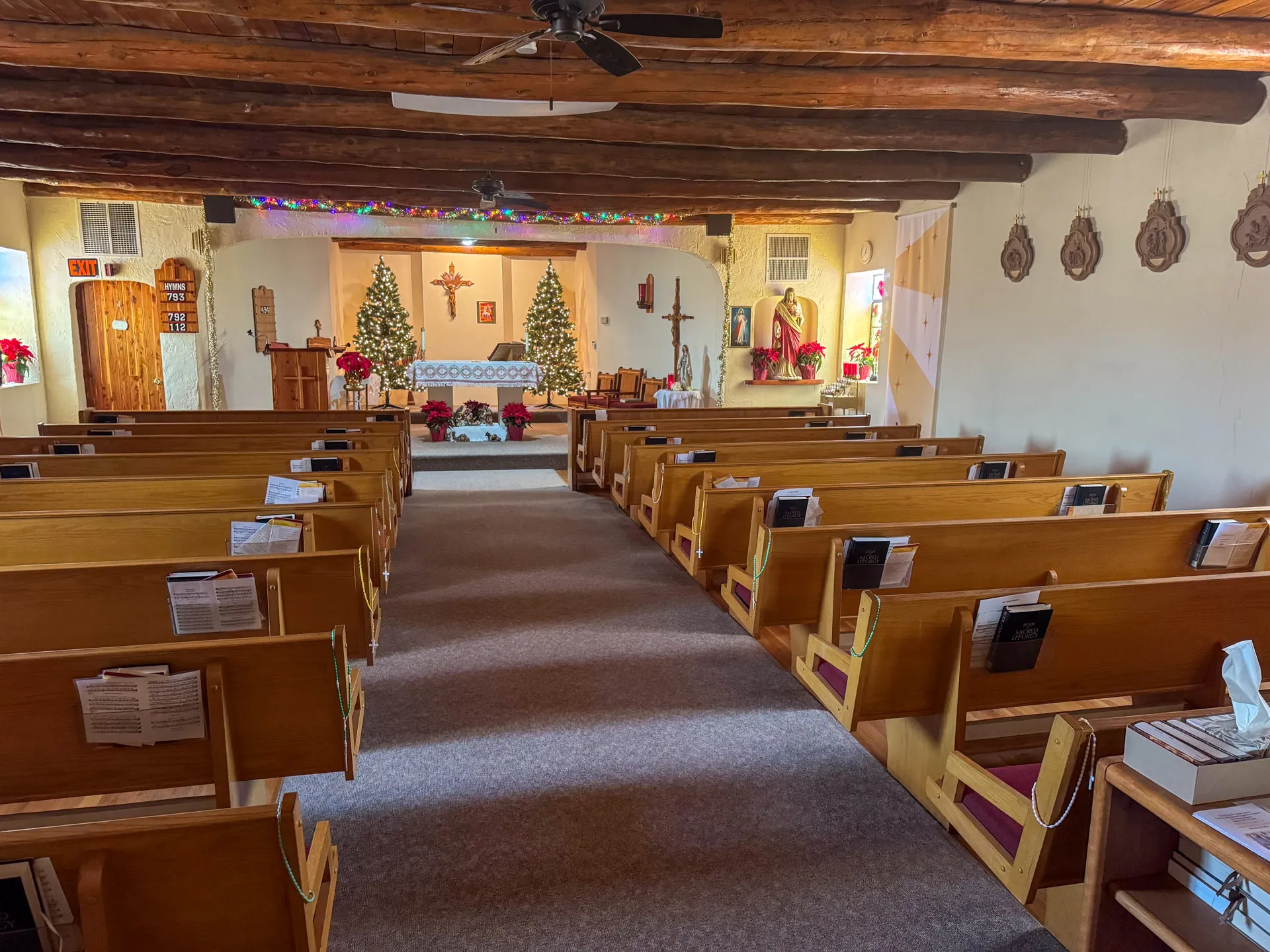 Interior of a church with wooden pews leading to an altar. The altar is adorned with two decorated Christmas trees and red poinsettias. There's a cross on the back wall, along with holiday garlands. Walls feature southwestern-style decor. A ceiling fan hangs from the wooden beam ceiling.