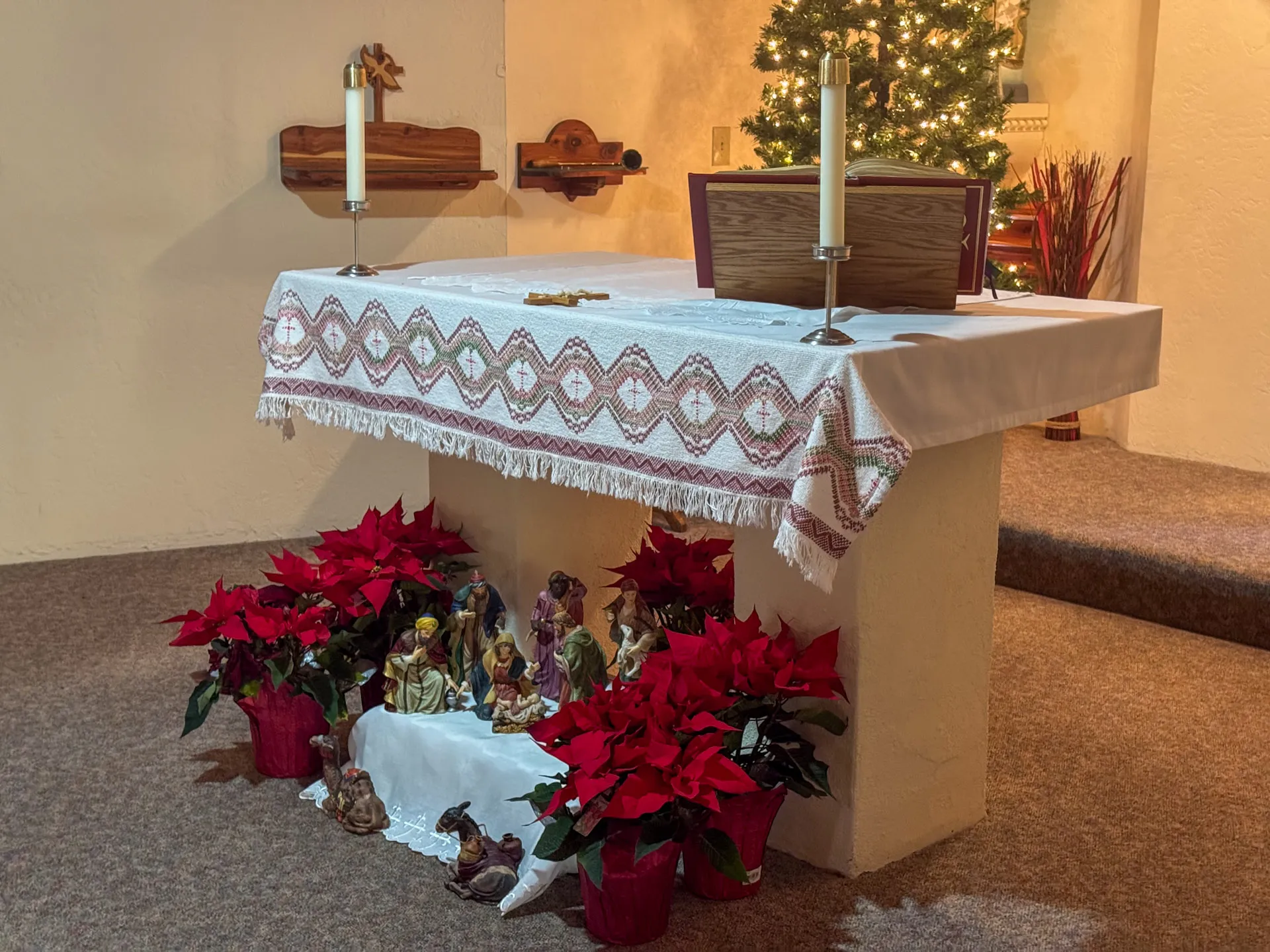 A church altar adorned with a white and red embroidered cloth is flanked by lit candles. Below, a Nativity scene with figures is surrounded by vibrant red poinsettias in pots. In the background, a decorated Christmas tree shines softly.
