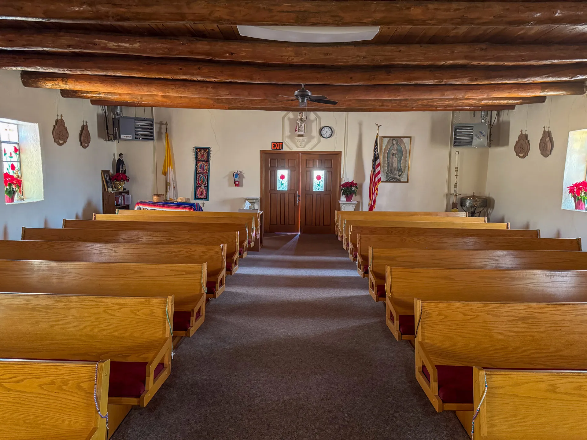 Interior of a church with wooden pews facing a wooden door at the back. The ceiling features exposed wooden beams. On the walls, there are small stained-glass windows, religious artwork, and a U.S. flag. Red poinsettias are placed near the altar area. A ceiling fan is centrally located.