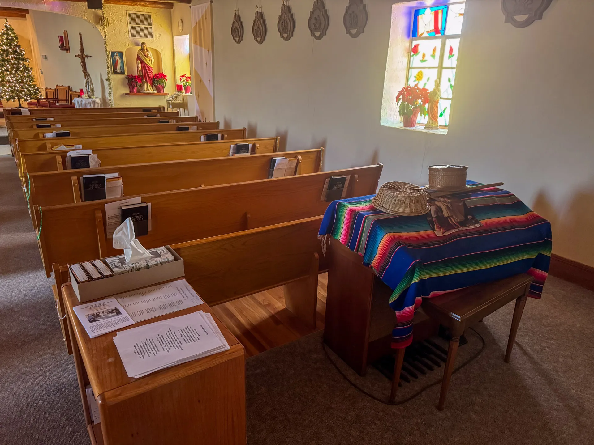 The image depicts the interior of a chapel with wooden pews, hymnals, and a lit Christmas tree in the background. A colorful striped cloth covers a small table with baskets. Light filters through a stained glass window, illuminating red flowers on the sill. Religious statues are present.