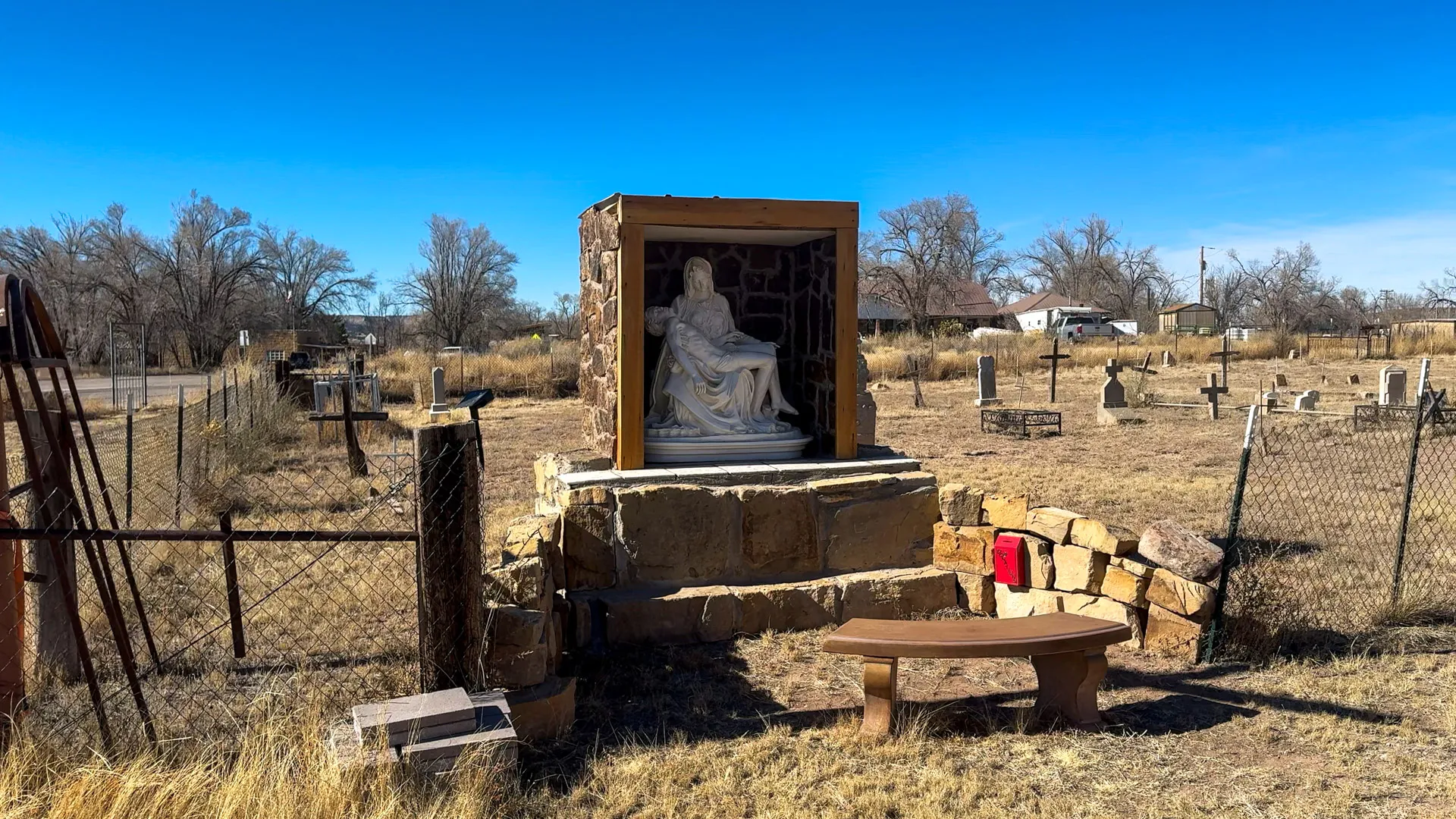 A serene cemetery scene features a stone sculpture of a seated figure within a wooden shelter. Surrounding the statue are crosses and headstones. A wooden bench and a red cloth rest on a stone barrier. The landscape is dry with sparse grass, and trees in the background under a clear blue sky.