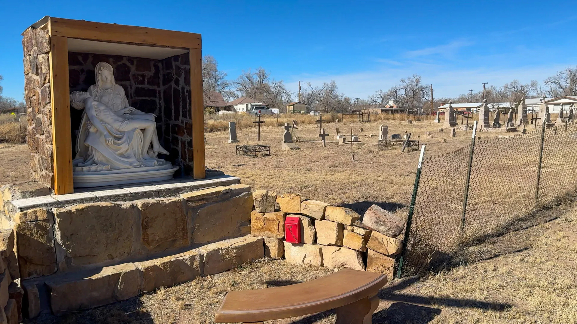 A cemetery scene under a clear blue sky features a stone display with a white statue resembling Michelangelo's Pietà, in a wood-framed niche. Nearby, a wire fence encloses the area, alongside a wooden bench and a small red box on a stone wall. Scattered headstones are visible in the background.