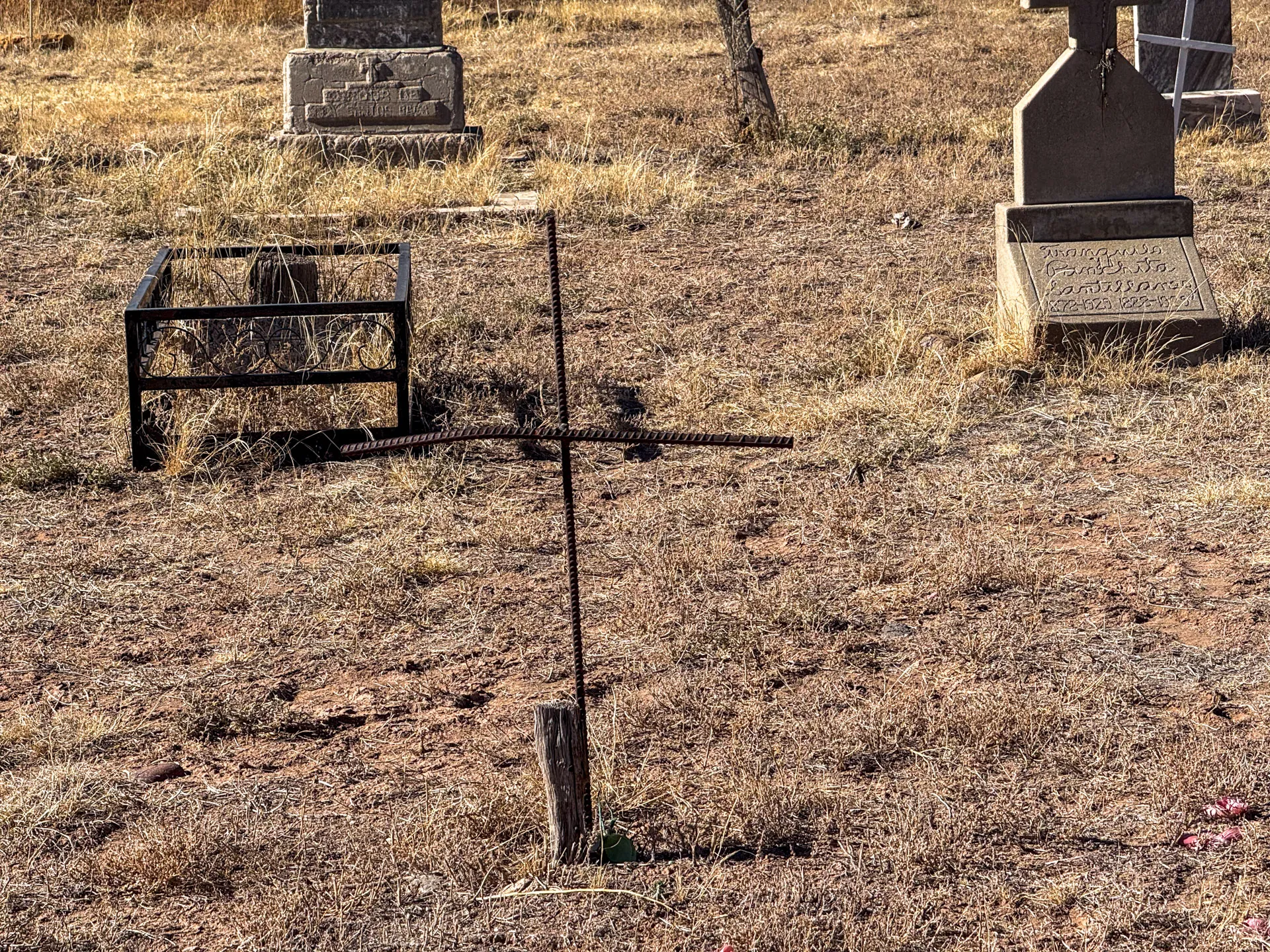 A rustic cemetery scene with a makeshift cross made of iron bars on a wooden base in the foreground. Surrounding the cross are dry grass and two weathered stone headstones. One headstone is partially legible, and the other is enclosed by a simple iron fence.