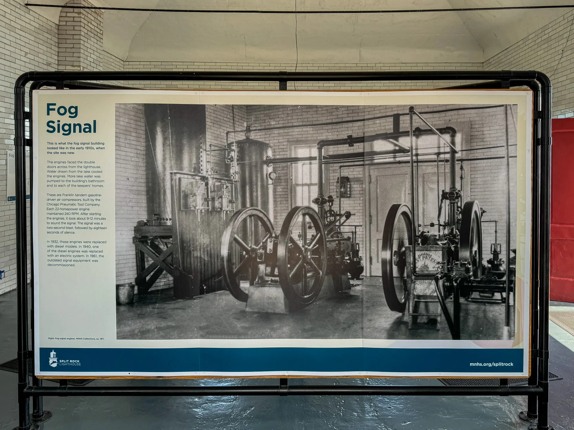 A sign titled "Fog Signal" displays a black-and-white photo of a historical maritime fog signal apparatus. It features large wheels, pipes, and a steam piston engine. The sign is mounted inside a brick building, with text explaining the equipment’s purpose and history.