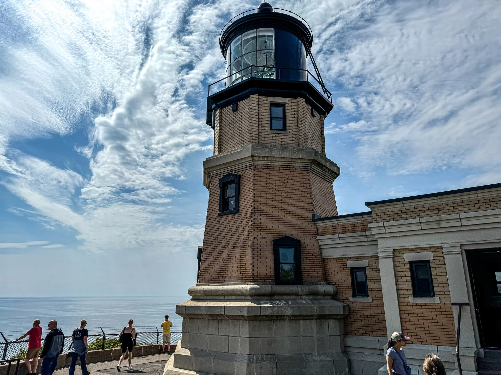 A tall brick lighthouse with a black metal top stands under a partly cloudy sky. It is next to a low building. The ocean is visible in the background, and several people are walking around the lighthouse area, with some looking out at the ocean.