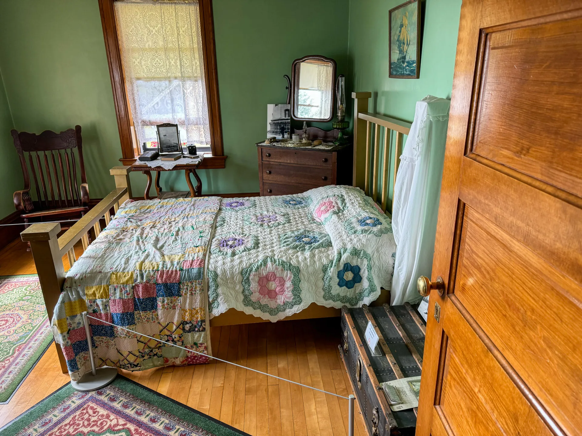 A cozy, vintage bedroom with green walls features a wooden bed adorned with a colorful patchwork quilt. A wooden dresser with a mirror holds various items. A rocking chair sits by a window with lace curtains. A trunk is at the bed's foot, and a patterned rug covers the floor.