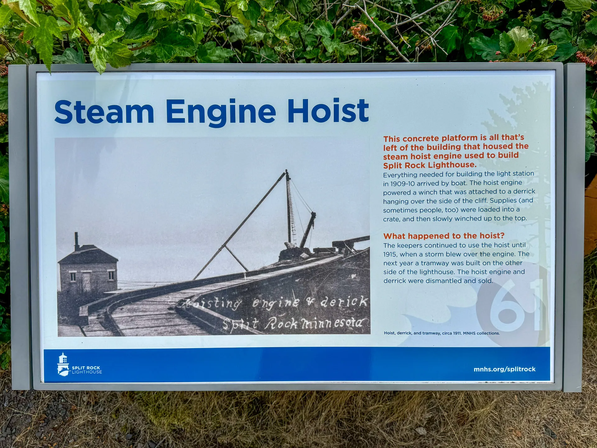 Sign explaining the Steam Engine Hoist at Split Rock Lighthouse. Includes a historic photo of a steam engine and derrick with text about its use in 1909 to transport materials for lighthouse construction. Background shows greenery and part of the old building's foundation.