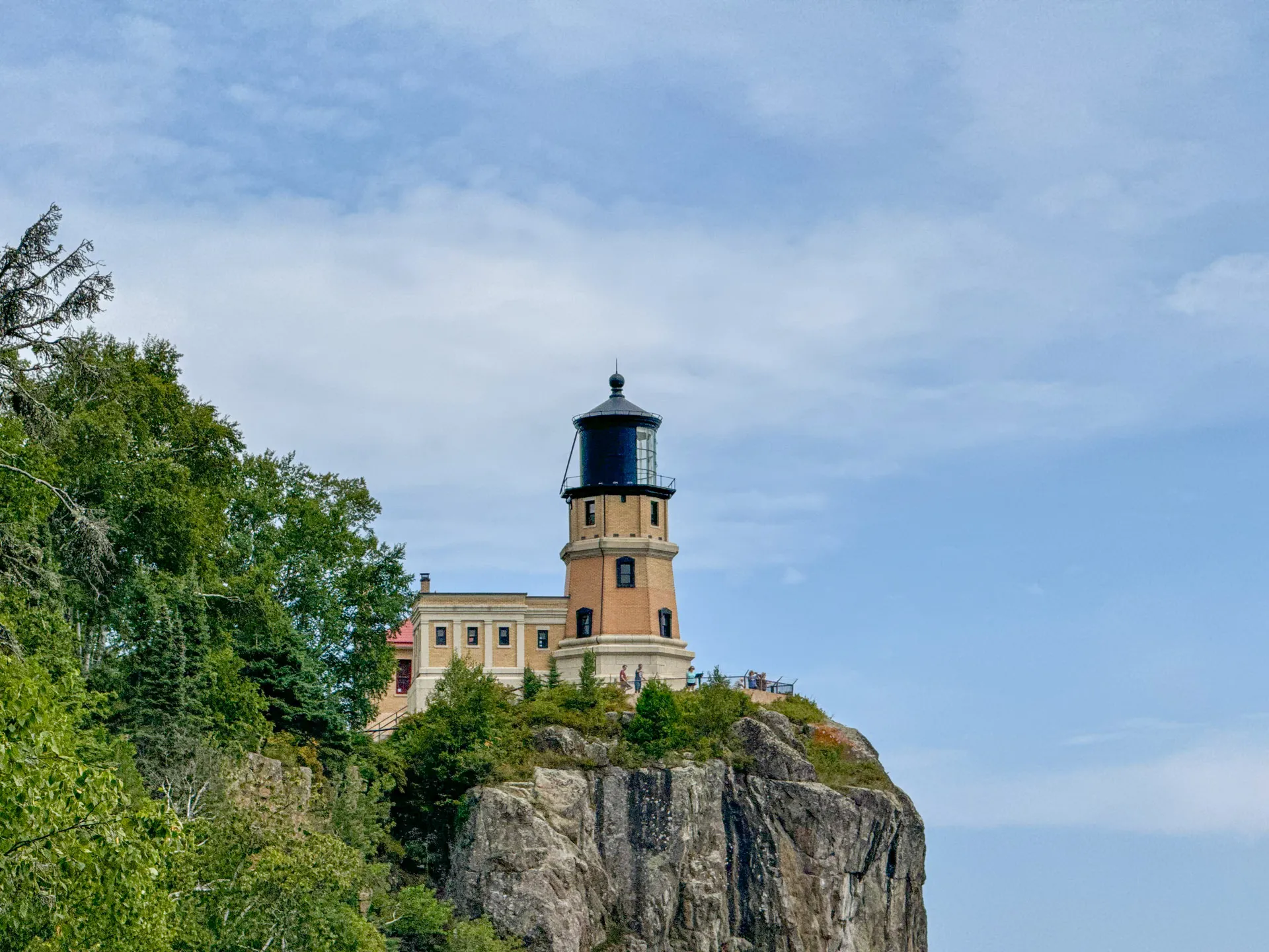 The image shows a lighthouse perched on a steep cliff. The structure is beige with a black top, surrounded by trees and greenery. A vast blue sky with few clouds serves as the backdrop, giving a serene and scenic atmosphere to the landscape.