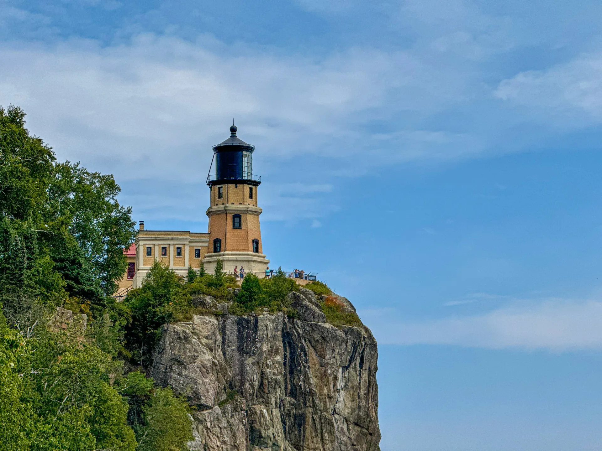 The image depicts a lighthouse perched on a rocky cliff with lush greenery at the base. The sky is mostly clear with a few scattered clouds. The lighthouse is cylindrical with a light at the top, positioned against a backdrop of blue sky and serene landscape.