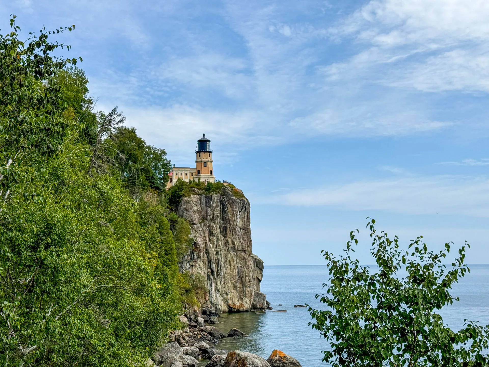 A lighthouse sits atop a rugged cliff overlooking a calm, expansive body of water. The cliff is surrounded by dense green foliage, with trees framing the scene. The sky is partly cloudy, casting soft light over the landscape, creating a serene and picturesque view.