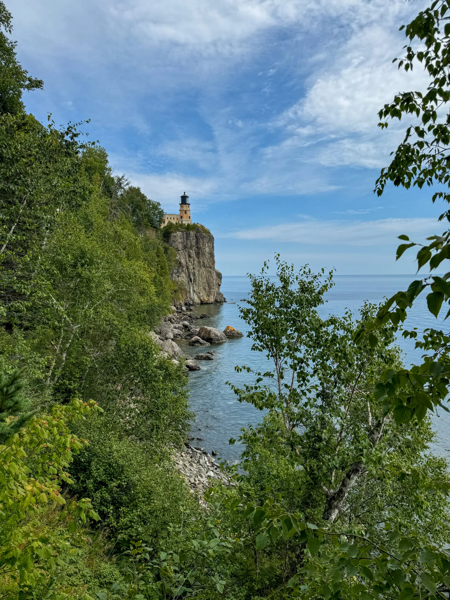 A lighthouse sits atop a steep rocky cliff overlooking a calm, expansive body of water. The sky is partly cloudy with patches of blue. Dense green foliage surrounds the area, framing the lighthouse and water, creating a serene, natural landscape.