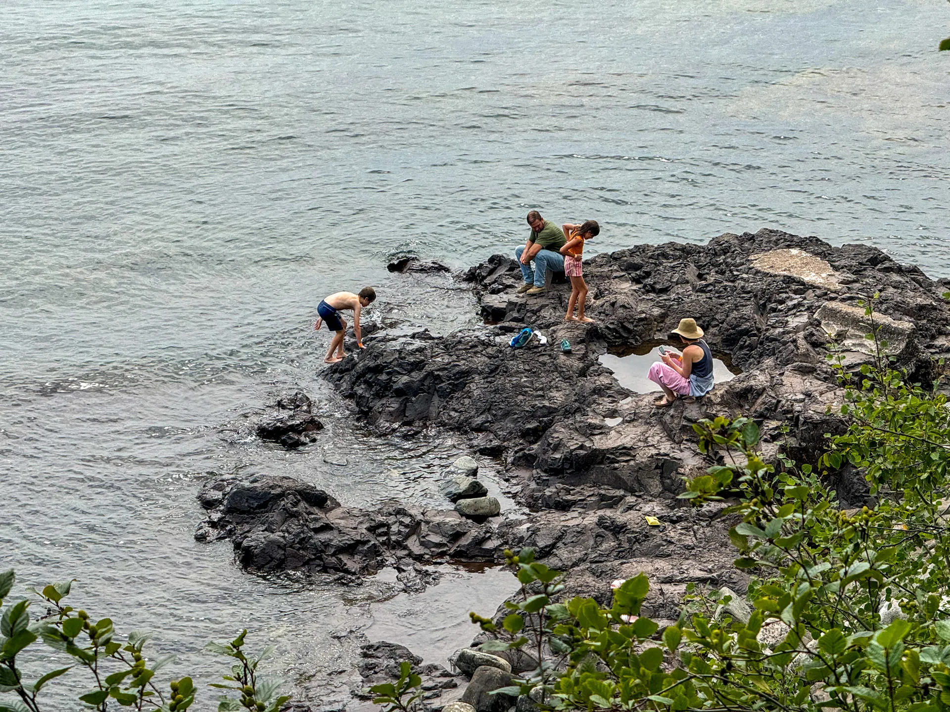 Four people explore rocky terrain by the ocean. Two individuals stand near the water's edge, and two sit on the rocks. The water is calm. Green foliage frames the foreground. The group appears relaxed, enjoying the natural setting.