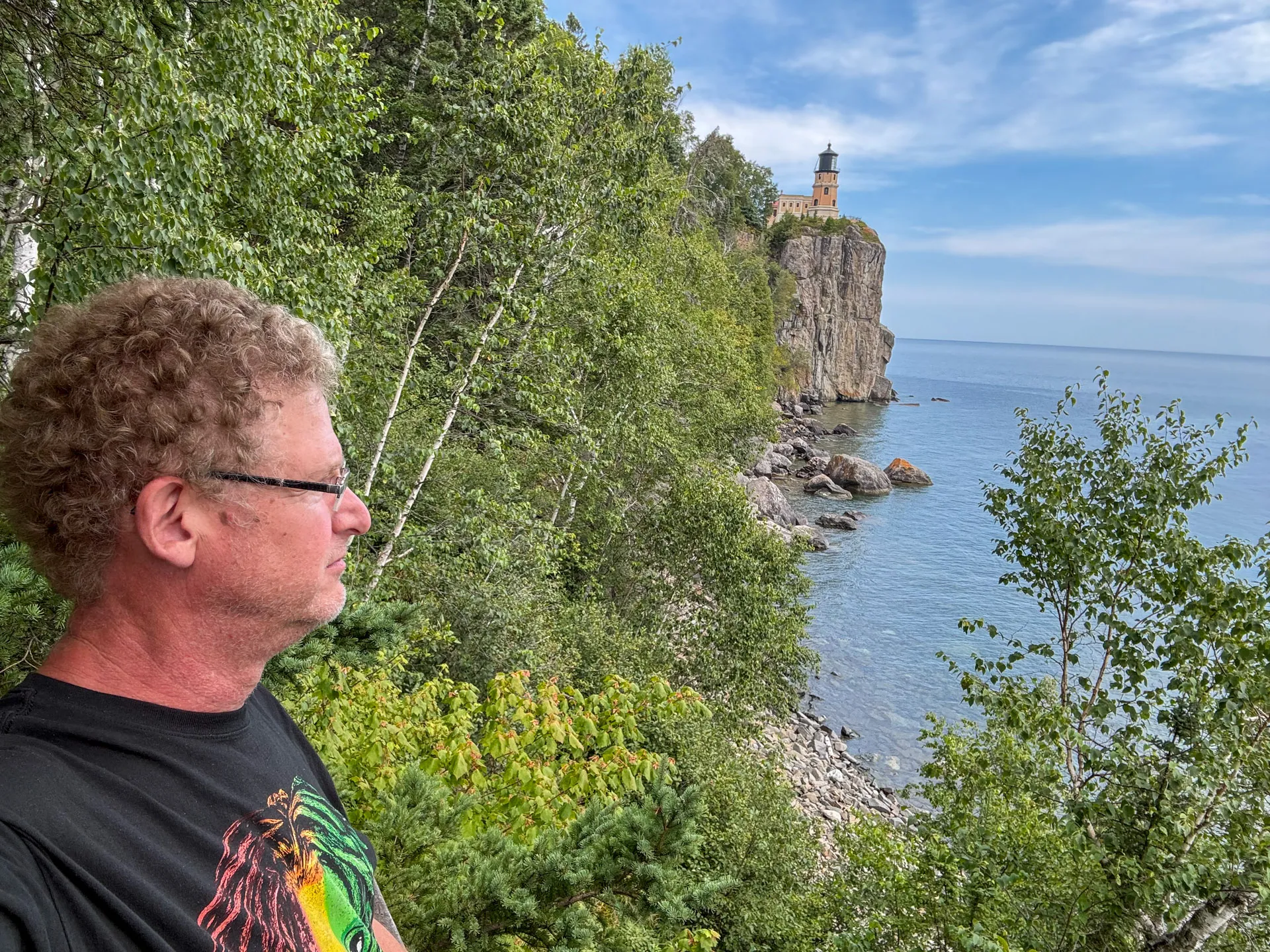 A man wearing glasses and a black shirt with a colorful graphic stands on a lush, tree-lined cliff overlooking a rocky shoreline. In the distance, a lighthouse stands atop another cliff. The sky is partly cloudy, and the ocean is calm, reflecting the blue of the sky.