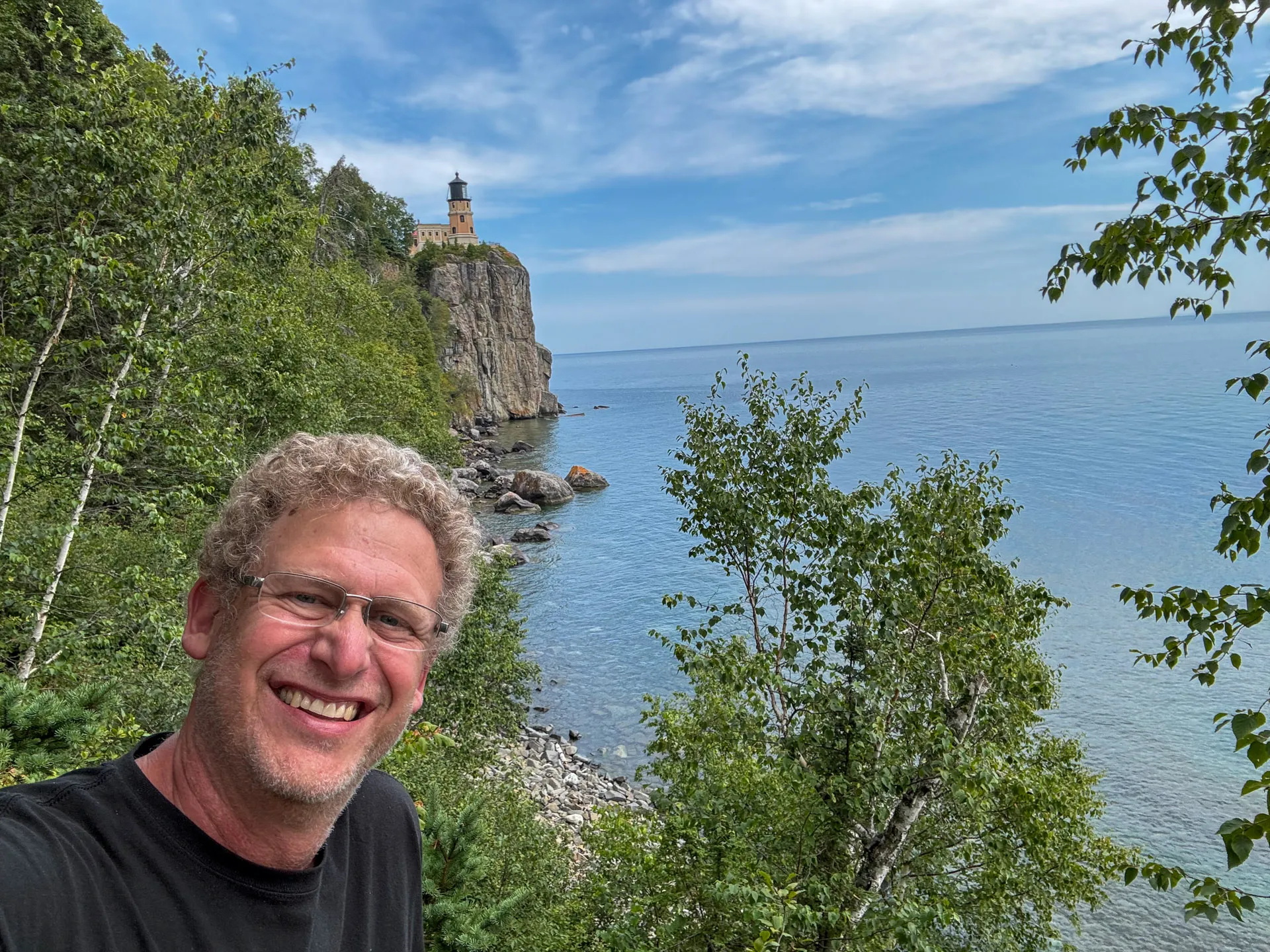 A person with curly hair, glasses, and a smile stands in the foreground. Behind them, a lighthouse sits on a cliff overlooking a large body of water. The scene is surrounded by lush green trees, and the sky is partly cloudy and blue.