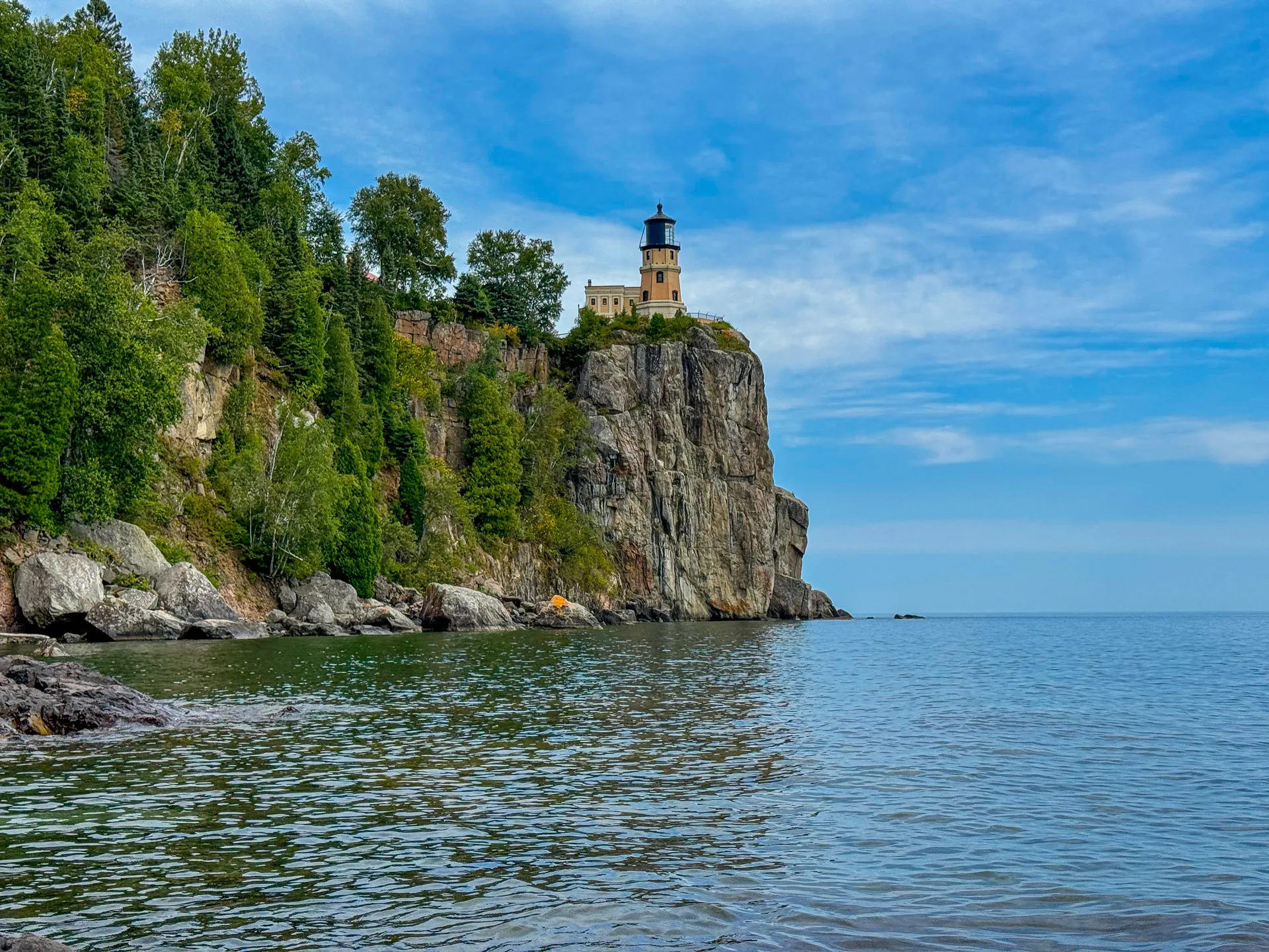 A lighthouse sits atop a rugged cliff surrounded by lush green trees, overlooking a calm, expansive body of water. The sky is mostly clear with wispy clouds. The rocky shoreline leads to the clear blue horizon, creating a serene coastal landscape.