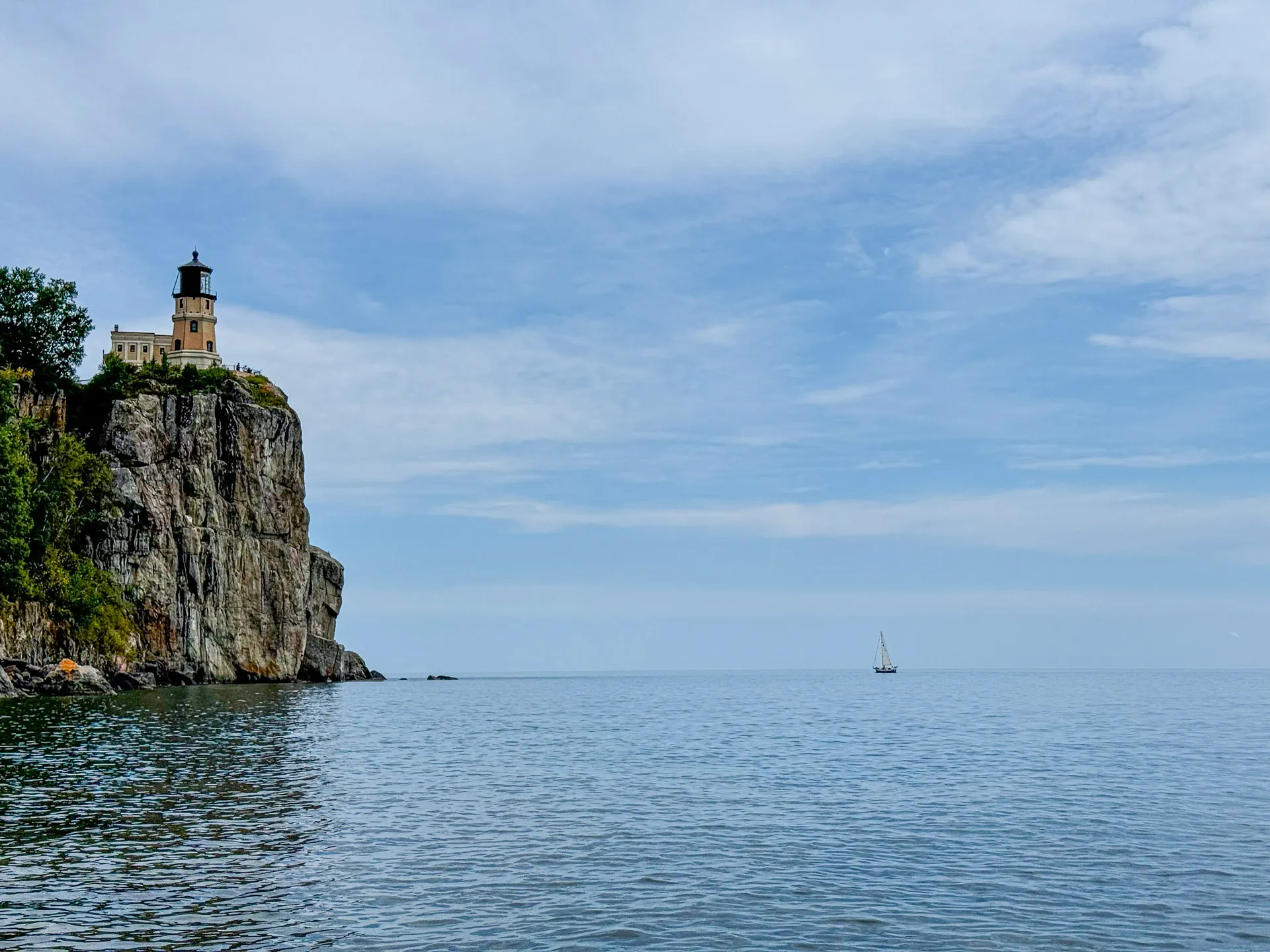 A lighthouse perches atop a rocky cliff on the left, surrounded by trees. Below, calm blue water stretches to the horizon, where a small sailboat is visible under a partly cloudy sky. The scene conveys a tranquil, coastal atmosphere.