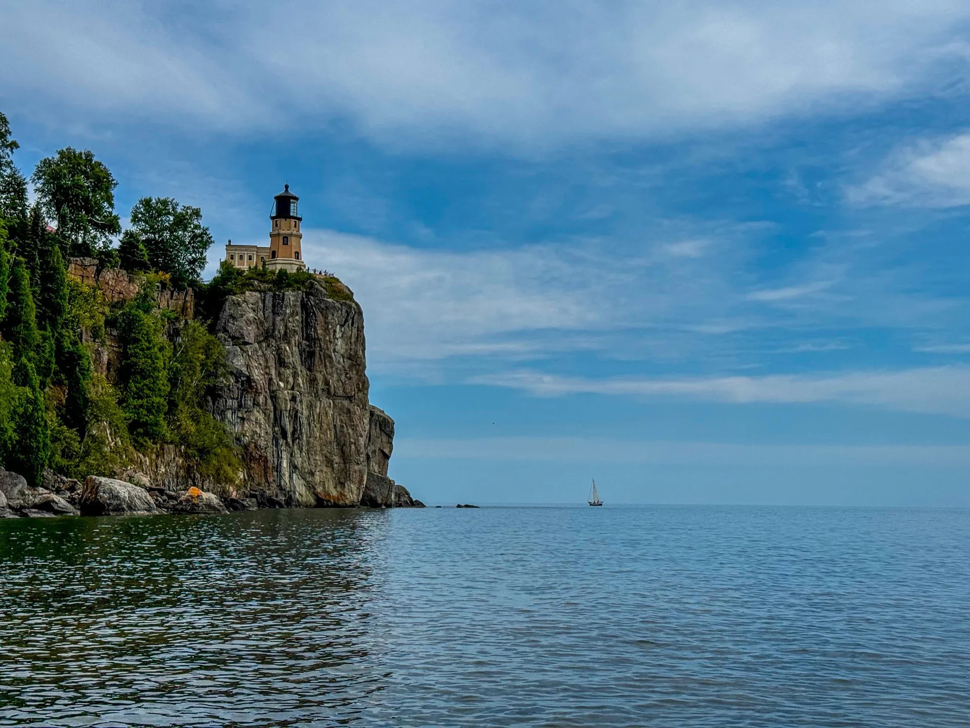 A lighthouse sits atop a steep, rocky cliff covered in greenery on the left. The vast, calm blue water of a lake extends to the right, where a small sailboat is visible in the distance. The sky above is partly cloudy with patches of blue.