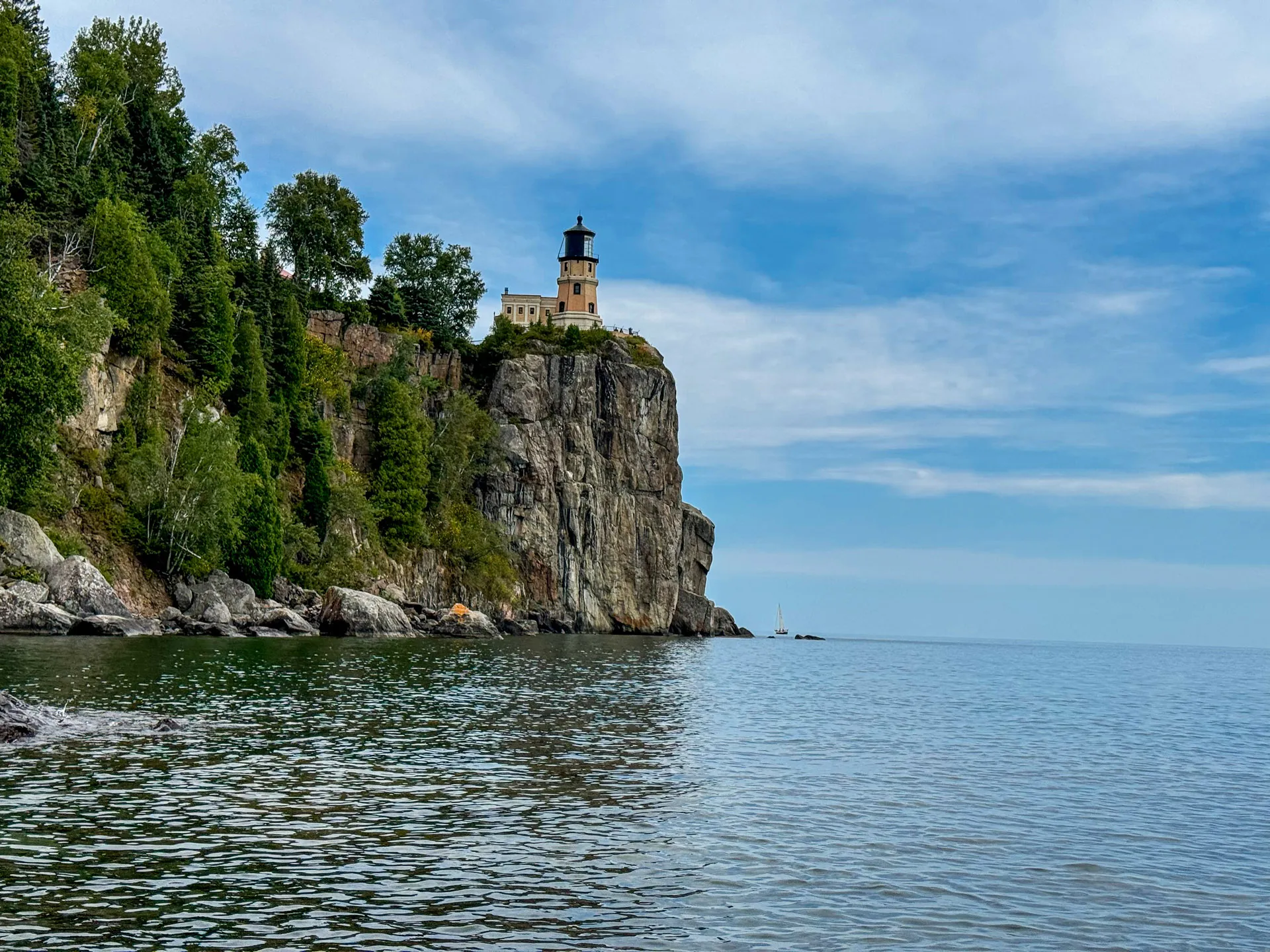 A lighthouse stands atop a steep, rocky cliff, surrounded by dense greenery. Below, calm waters stretch out, reflecting the blue sky dotted with clouds. In the distance, a sailboat is visible, adding to the serene coastal scene.