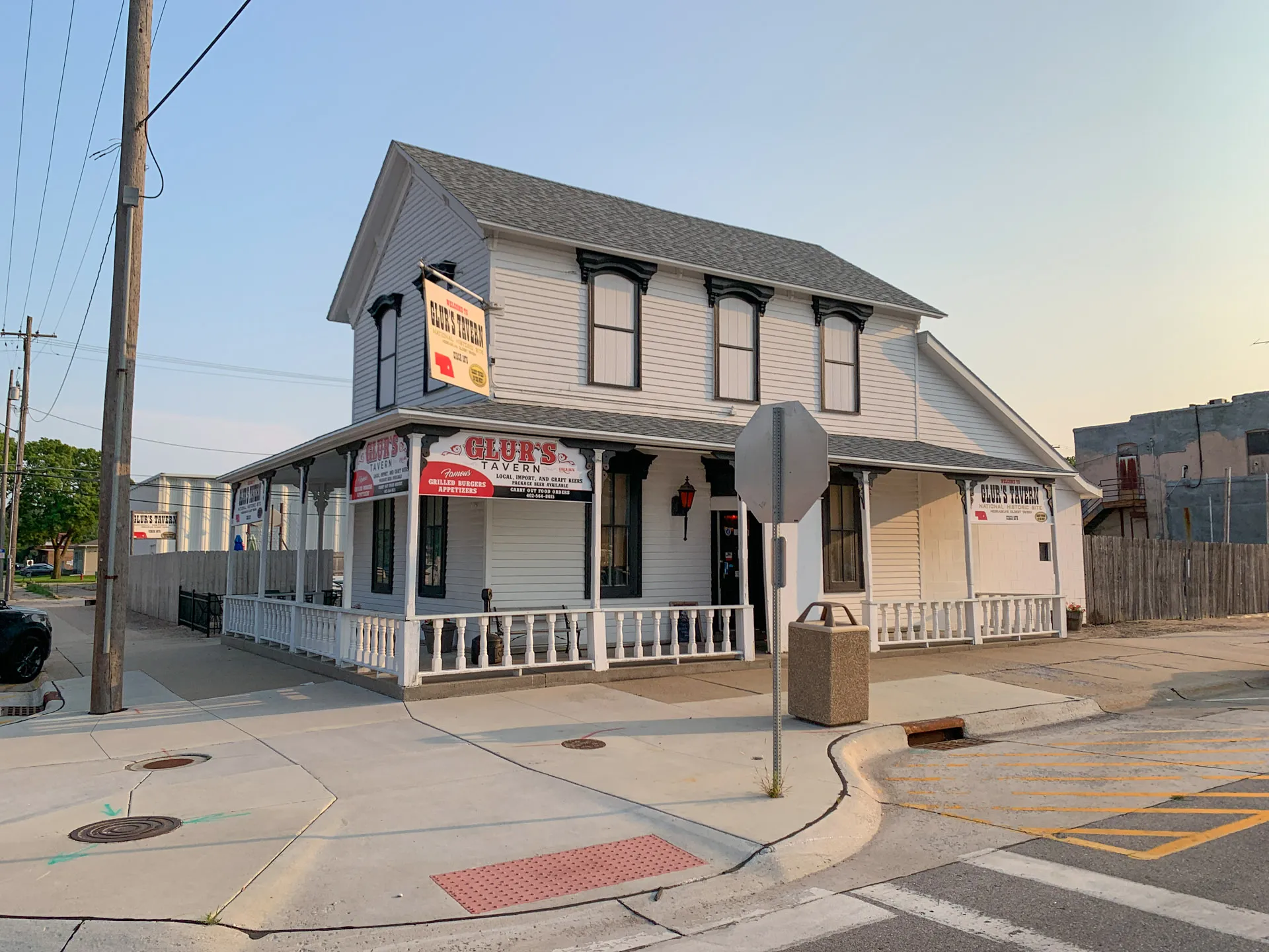A white, two-story building with gray roofing and an L-shaped porch, identified as a tavern. It has a "Glur's Tavern" banner and signage indicating food availability. The structure is adjacent to a street corner with a stop sign and concrete pavement. There's a chain-link fence and power lines nearby.