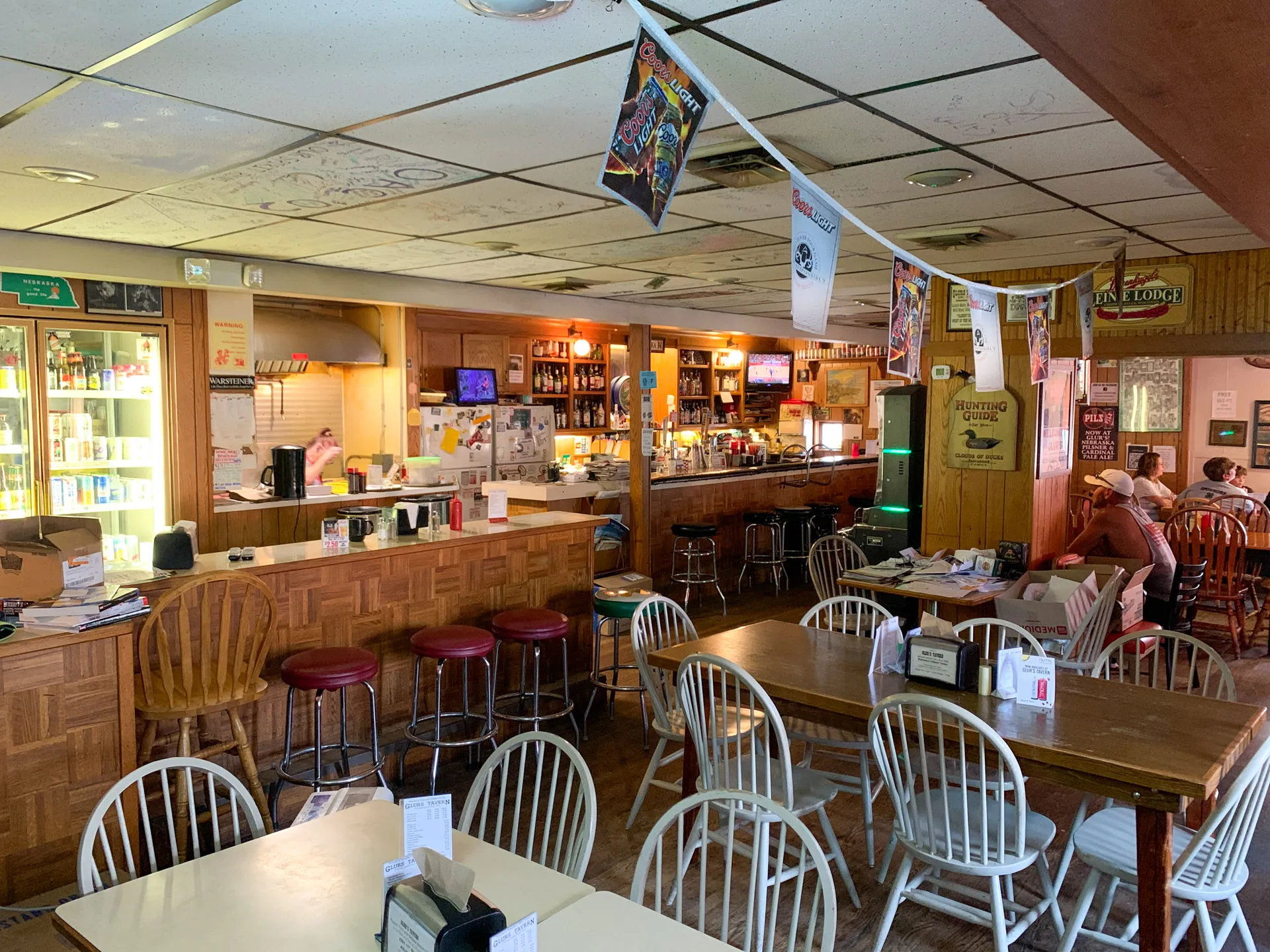 Cozy bar interior with wooden tables and chairs. The counter has red stools, bottles, and a cash register. Flags and posters hang from the ceiling. A fridge with drinks is on the left. Natural light streams in, and a few patrons sit in the background.