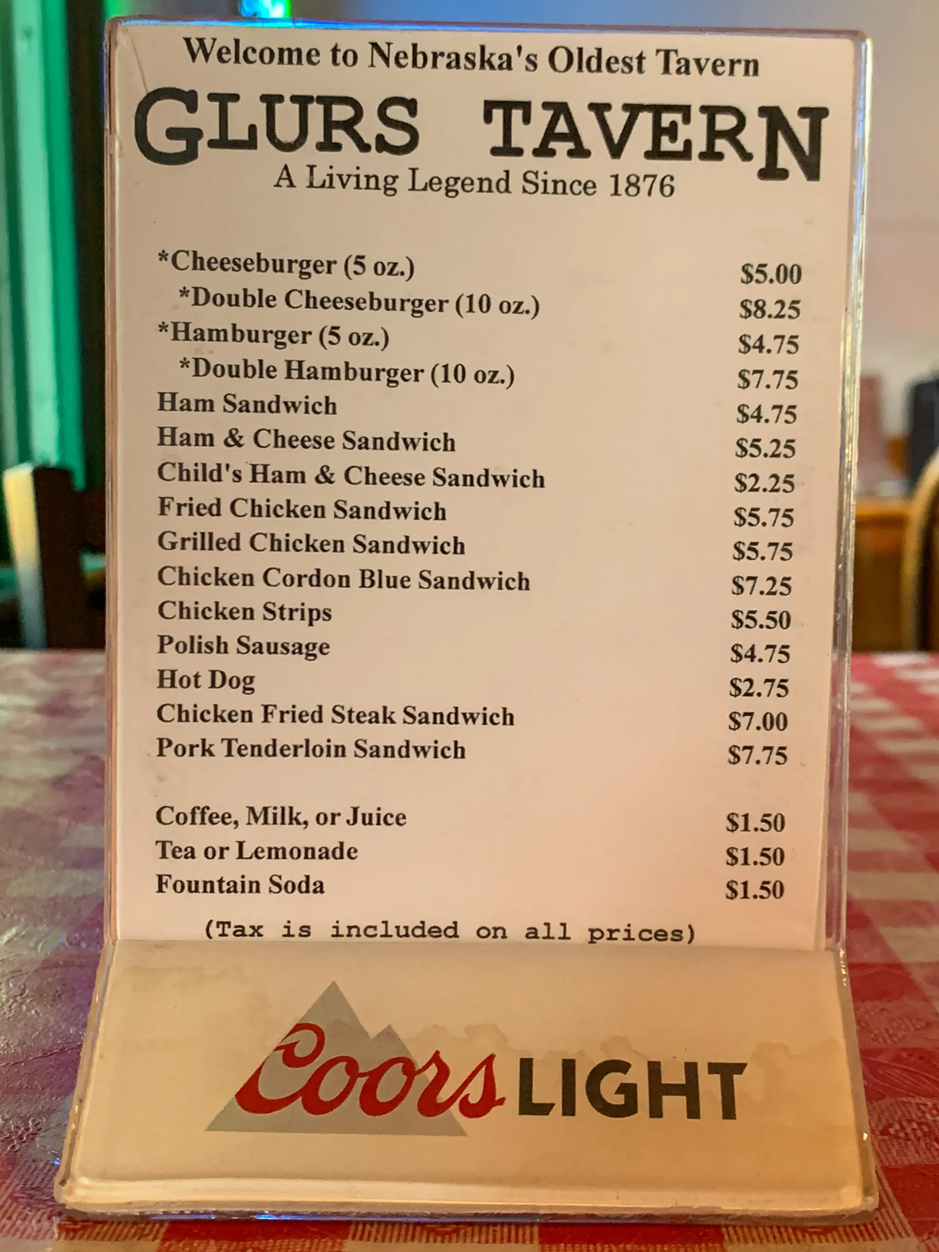Menu on a table with "Welcome to Nebraska’s Oldest Tavern" at the top, featuring items like cheeseburgers, hamburgers, sandwiches, and hot dogs, priced between $3.25 and $7.75. Drinks include coffee, milk, juice, tea, and lemonade. Coors Light logo displayed at the bottom.